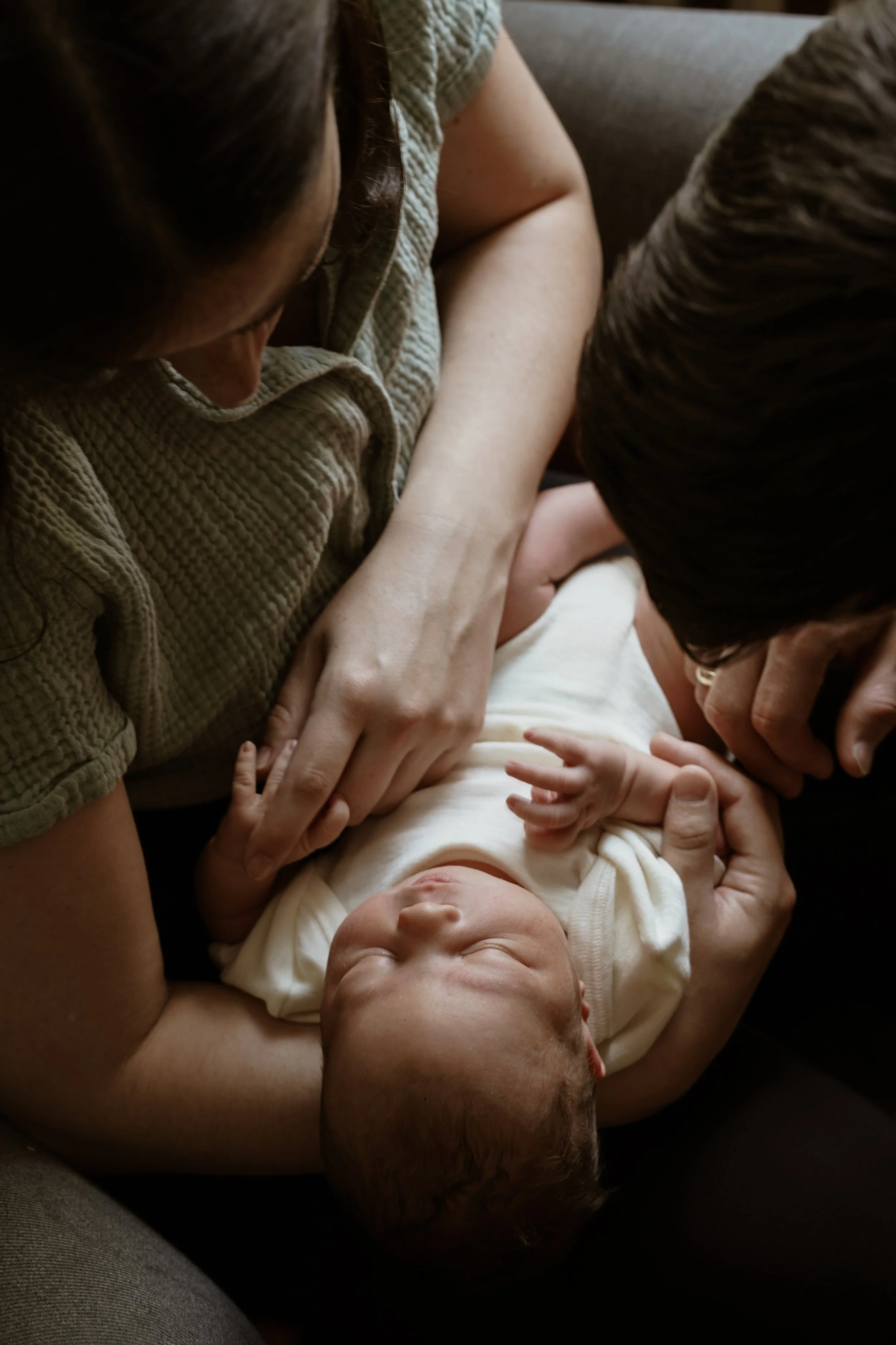 An adult laying on a sofa holding a baby who is lying on their back, smiling with eyes closed, wearing a white shirt, with another adult leaning over and touching the baby's shoulder.