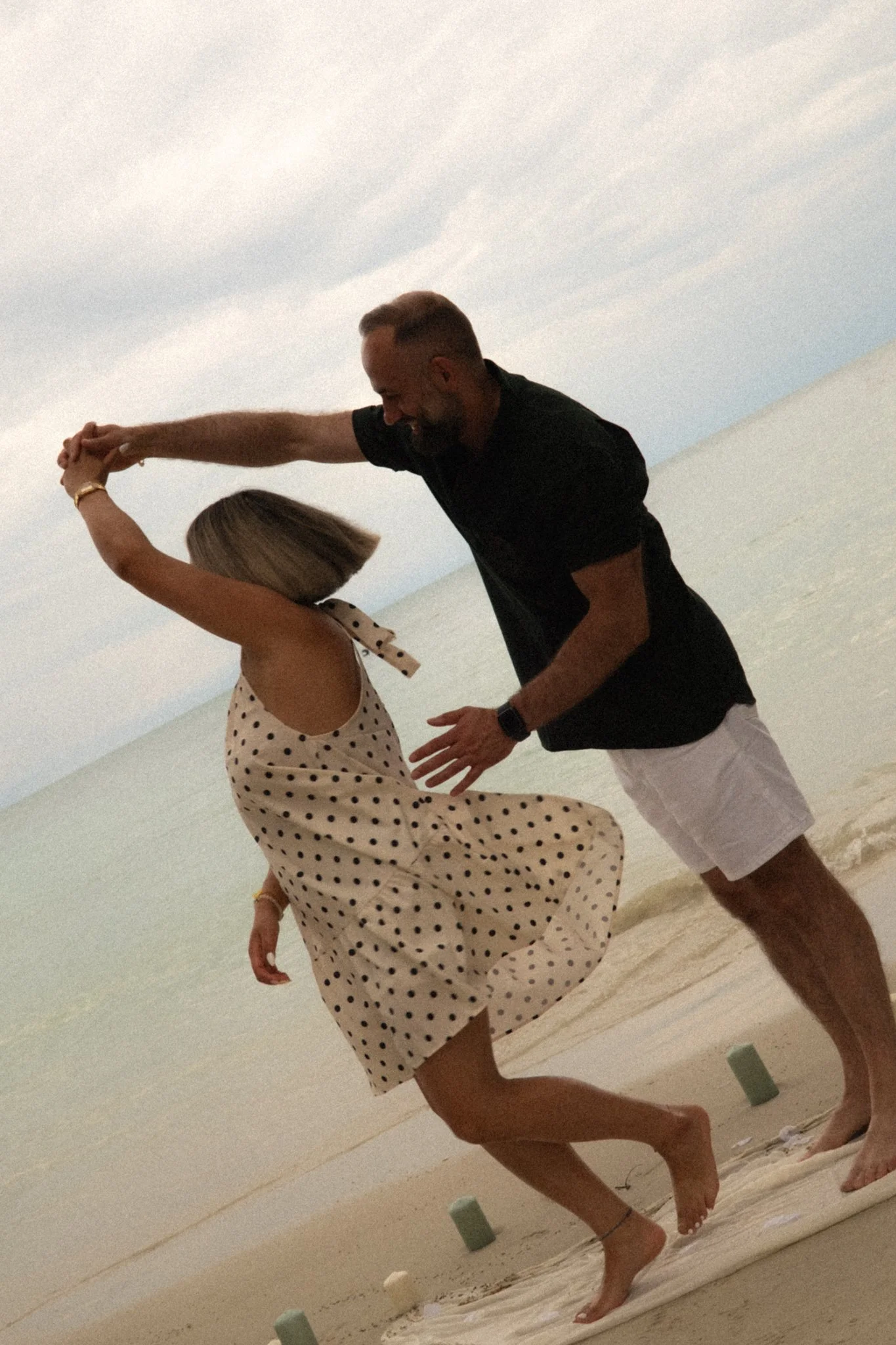 A man and a woman are dancing on a sandy beach, holding hands and twirling together. The woman is wearing a polka dot dress, while the man is dressed in a black t-shirt and white shorts. The sky is cloudy, and there are small cushions or objects scat