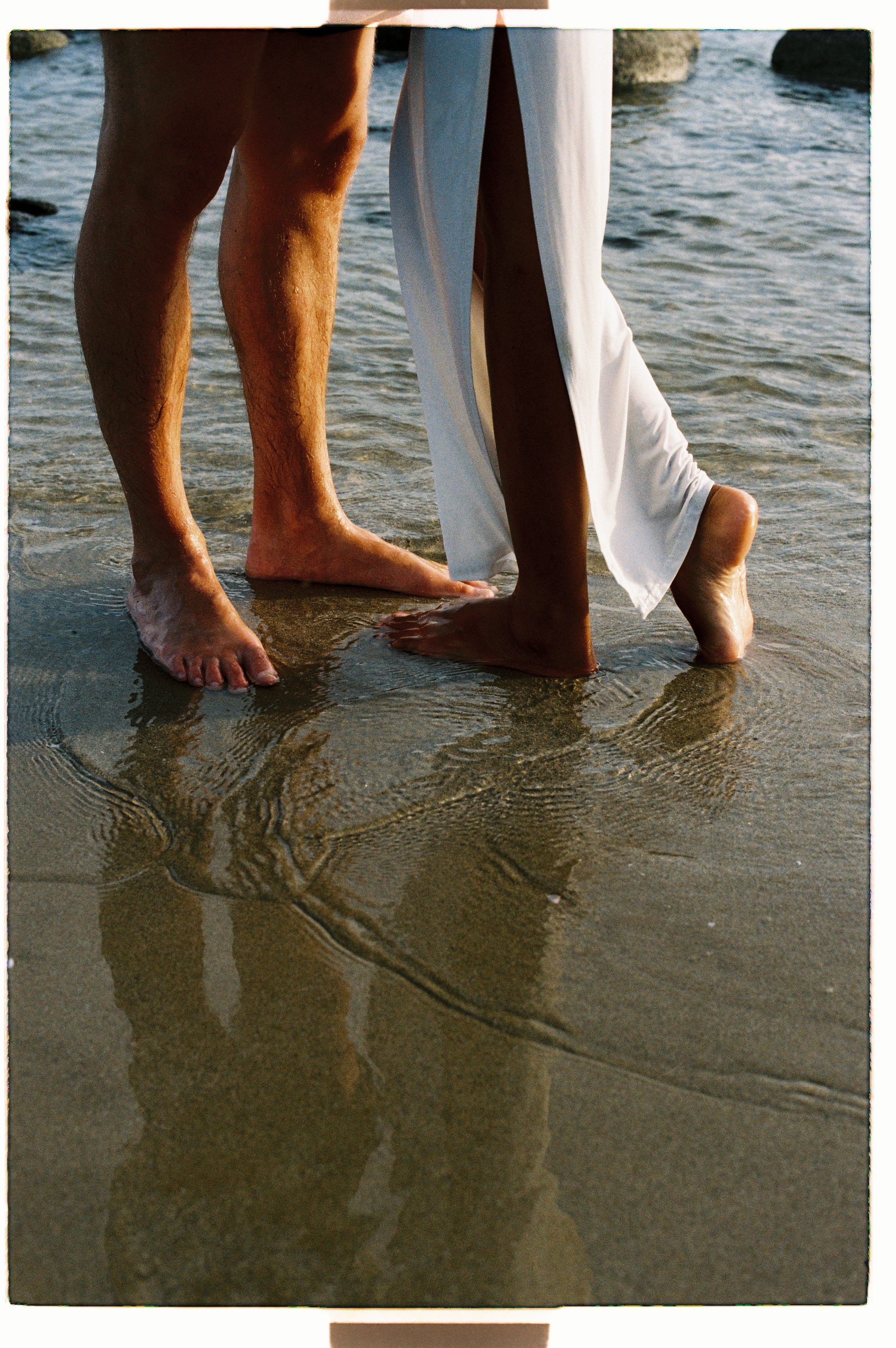 Close-up of two people standing in shallow water at the beach, one wearing white pants and the other with bare legs. Their reflections are visible on the wet sand.