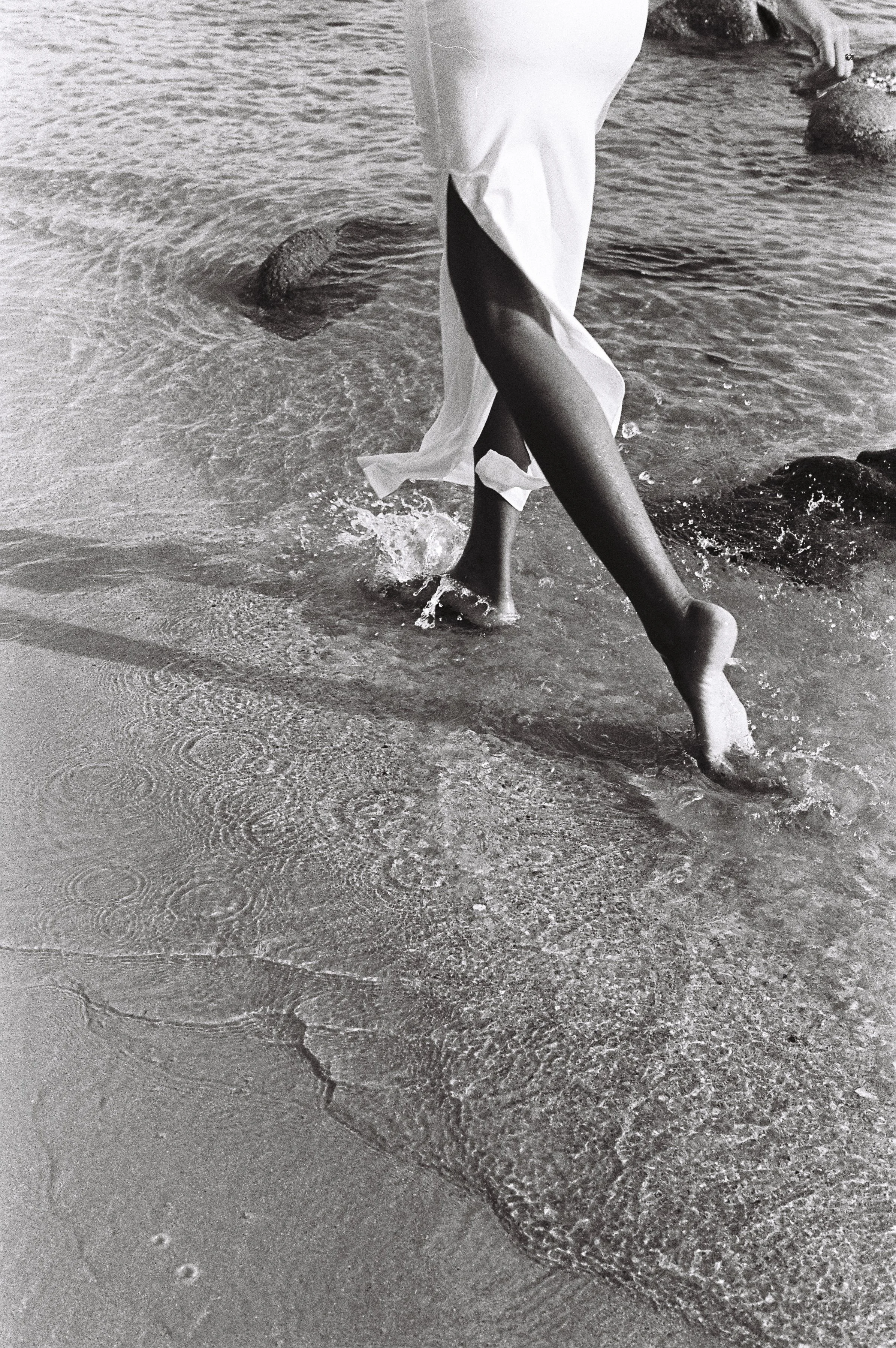 Person walking barefoot in shallow water at the beach, wearing a white dress.