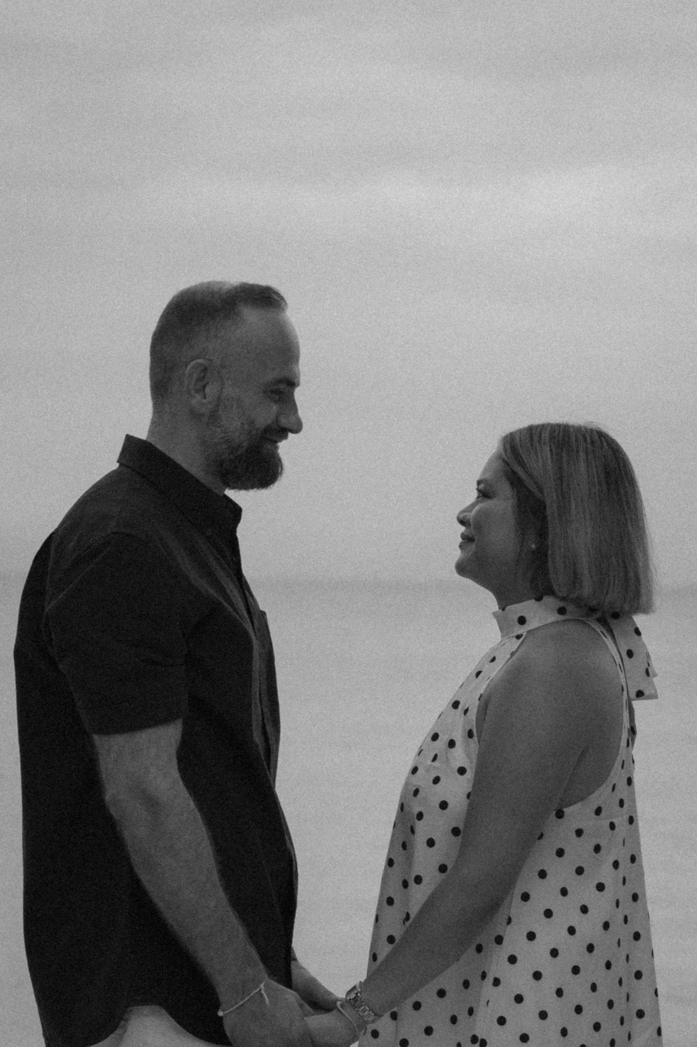 A man and a woman hold hands and look at each other, smiling, while standing outdoors against a cloudy sky.
