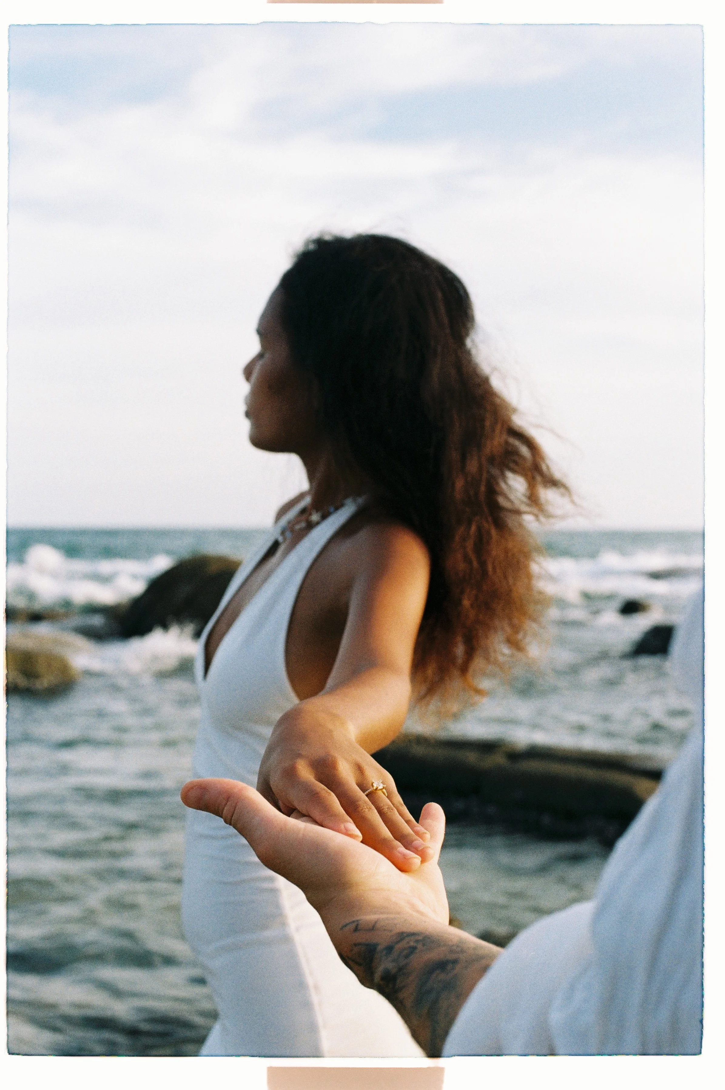 A woman in a white dress is at the beach, with waves and rocks in the background, standing with arms outstretched towards another person whose hand is holding her hand.