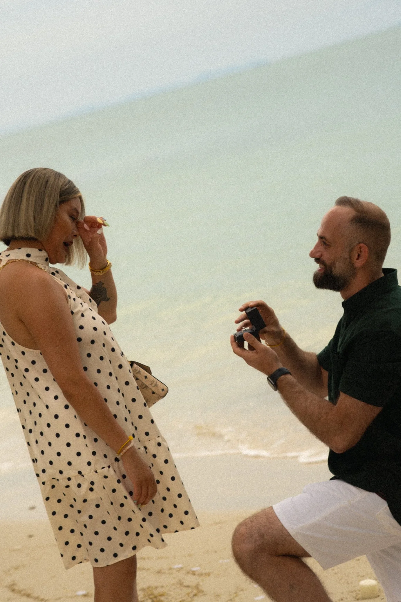A man proposing marriage to a woman on the beach, with an engagement ring box in his hand, woman appears surprised and emotional, sandy beach background.