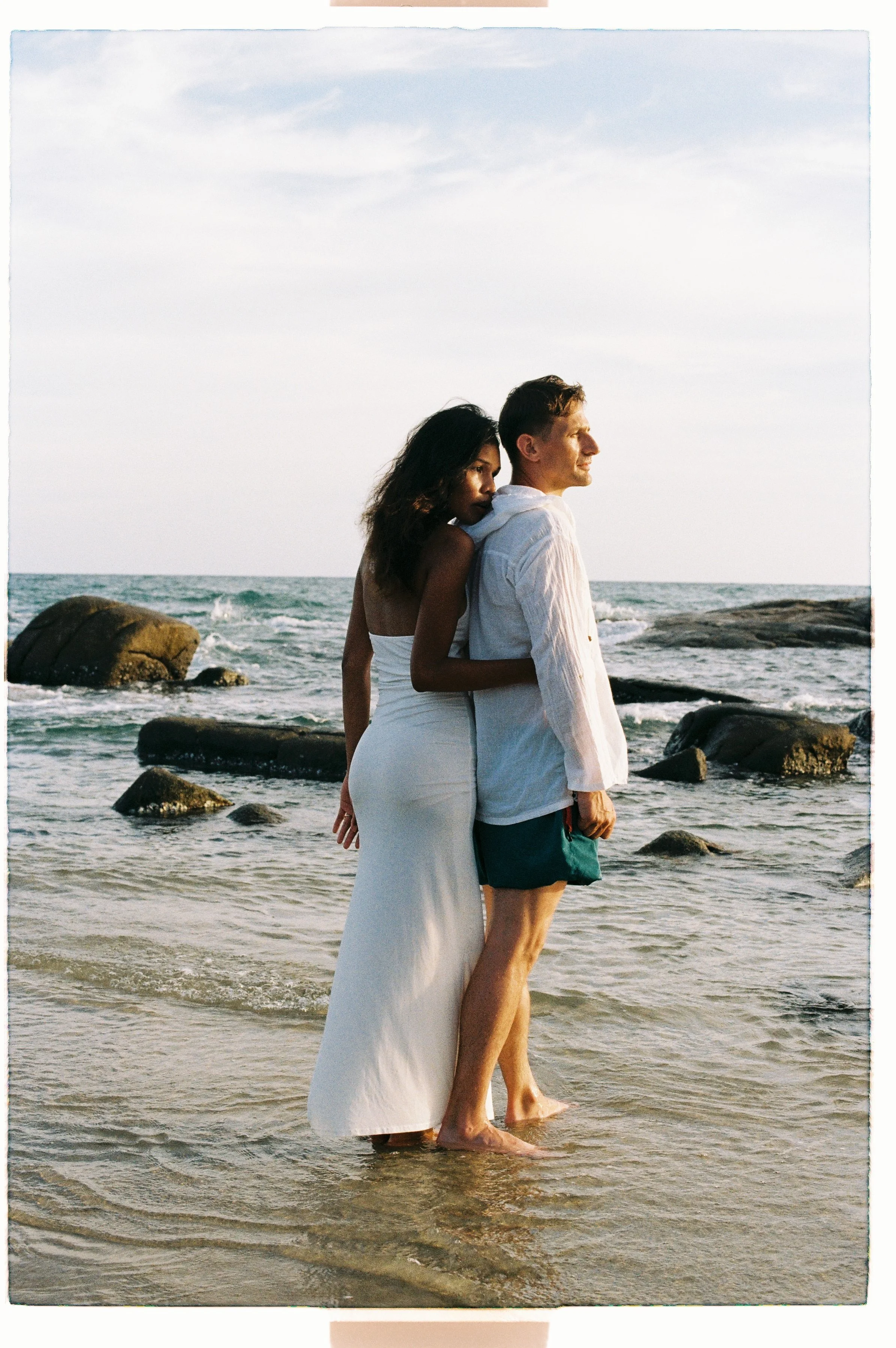 A couple standing close together on the beach near the water, with rocks and the ocean in the background, during sunset or sunrise.
