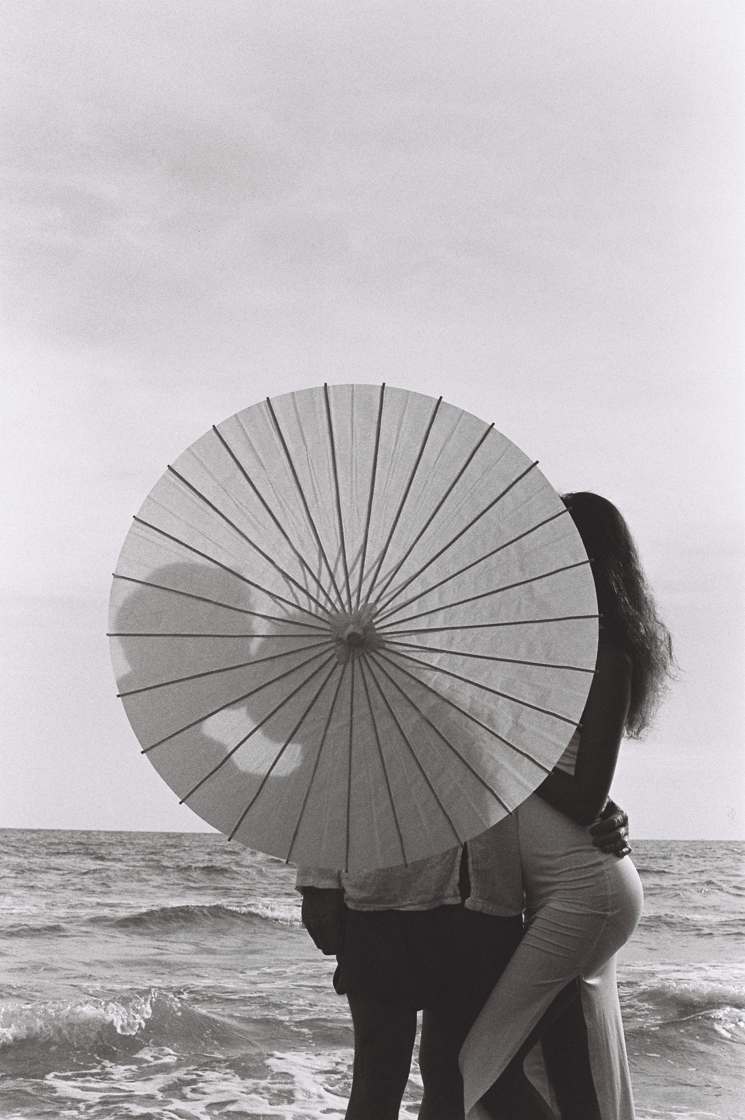 A couple standing on the beach, with the woman leaning into the man, holding an umbrella, during daytime. The ocean waves are visible at their feet, and the sky appears cloudy.