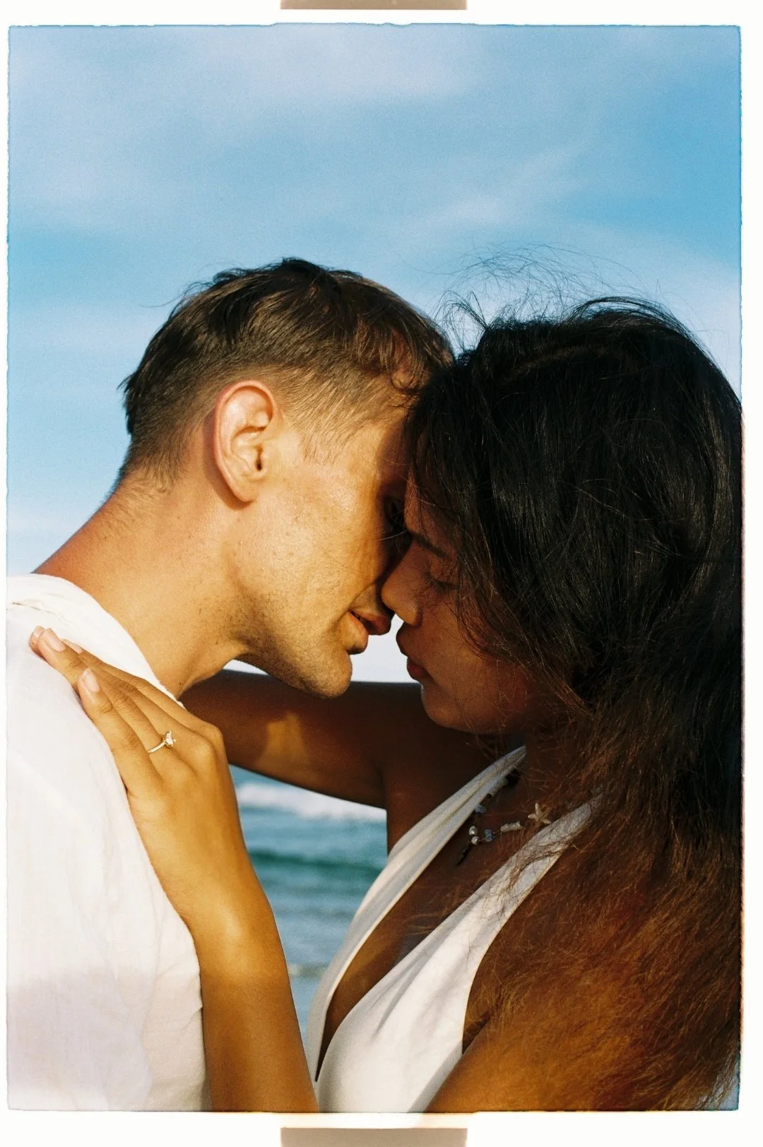 A man and woman with foreheads touching, eyes closed, embracing on a beach with ocean and blue sky in the background.