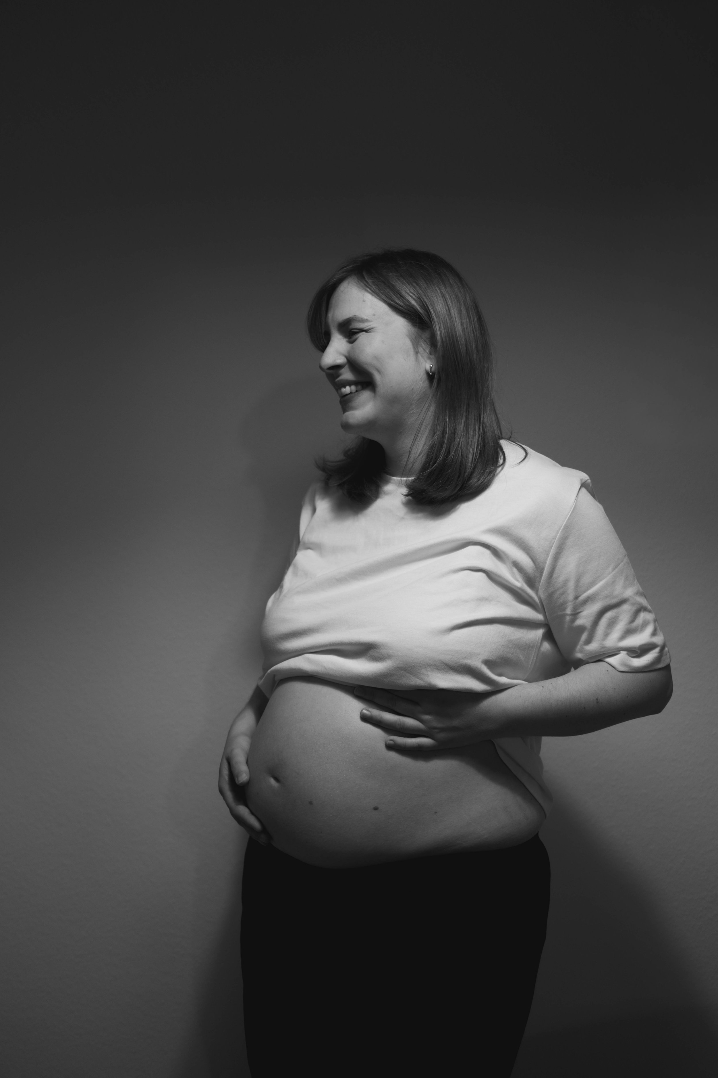 Black and white photo of a pregnant woman smiling, with her right hand on her belly and her left hand on her hip, standing against a plain background.