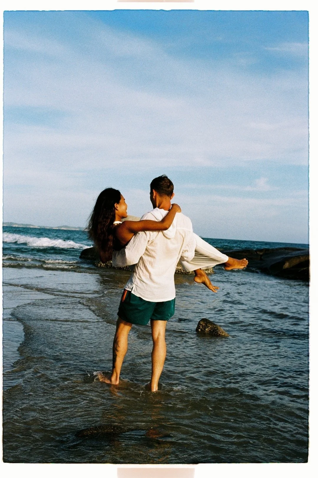 A man holding a woman in a white outfit while standing in shallow ocean water on a beach with rocks and a blue sky.