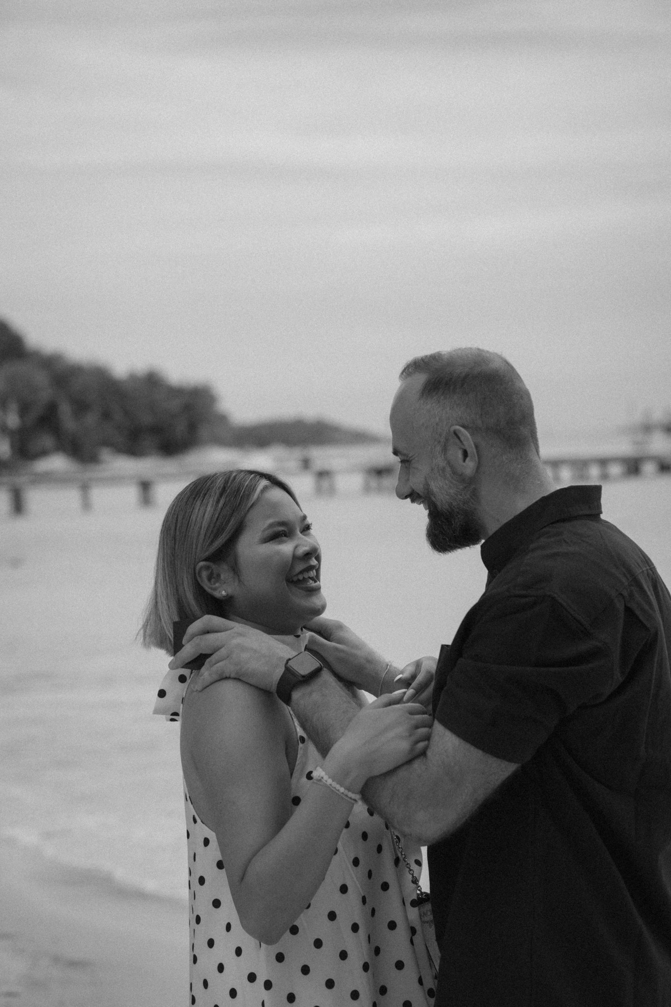 A smiling woman and a man are dancing closely on a beach, looking at each other joyfully with their hands gently touching. Behind them, there is a blurred view of the ocean, a sandy shore, and a bridge in the distance.