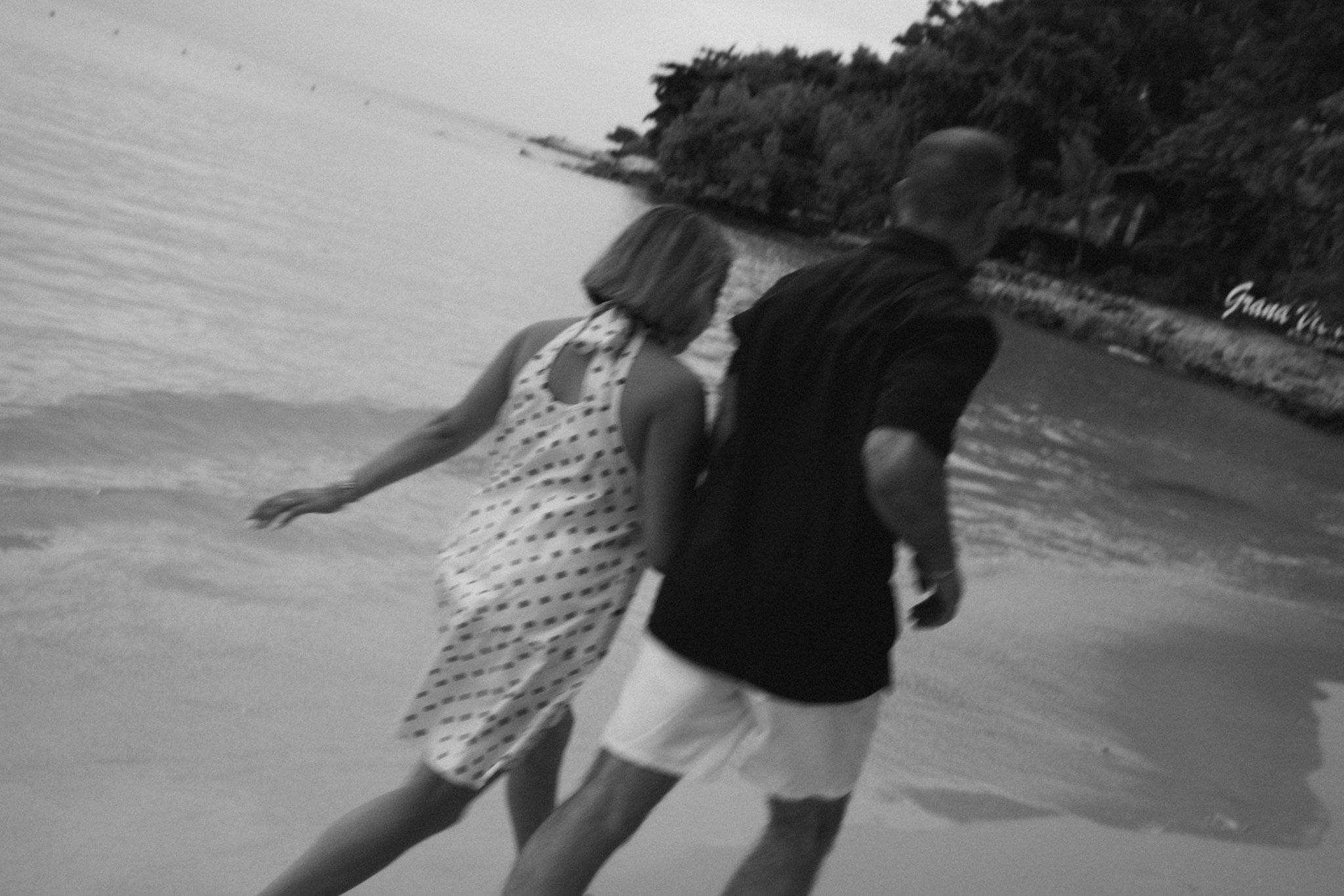 A black and white photo of two people walking on a beach, one woman wearing a polka dot dress and a man in casual clothes, with trees and a building in the background.
