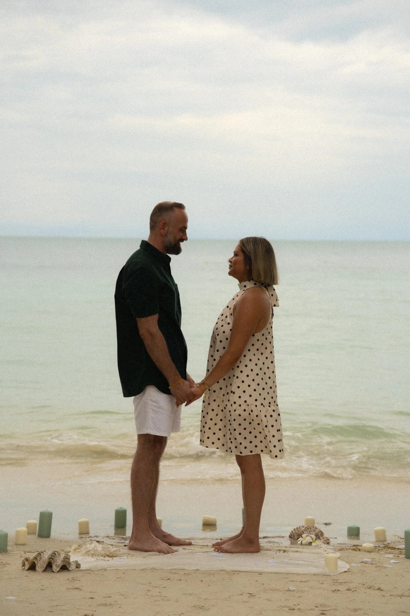 A couple stands face to face on a beach, holding hands and gazing into each other's eyes, with the ocean in the background.
