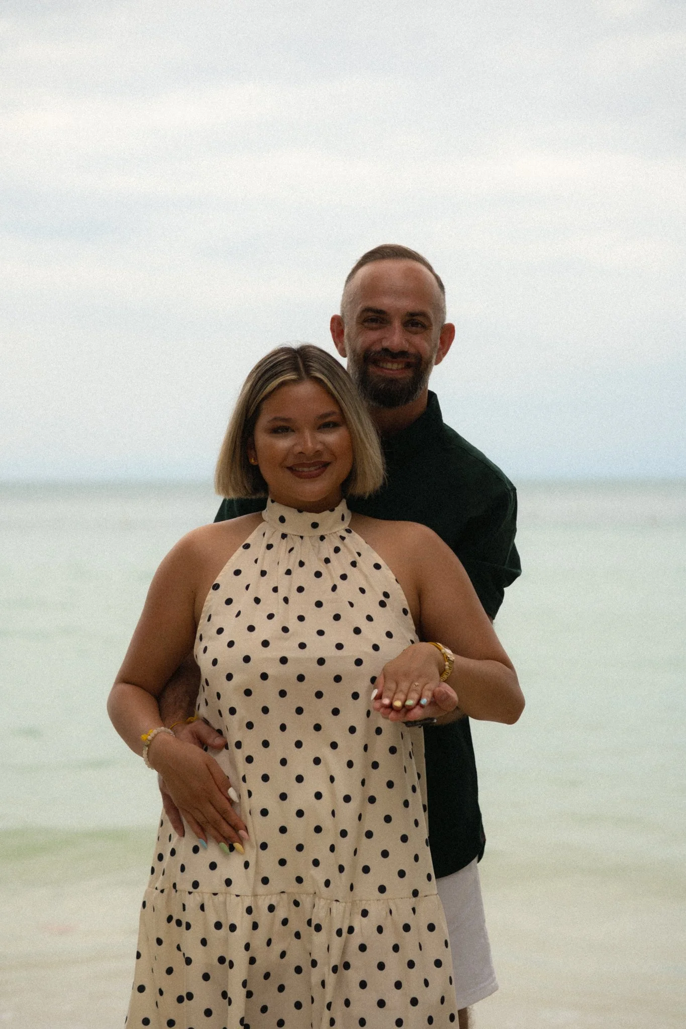 A couple standing close together on the beach, with the ocean and a cloudy sky in the background. The woman has blonde hair, is wearing a polka-dot dress, and has multicolored nails. The man has short hair, a beard, and is wearing a black shirt. They