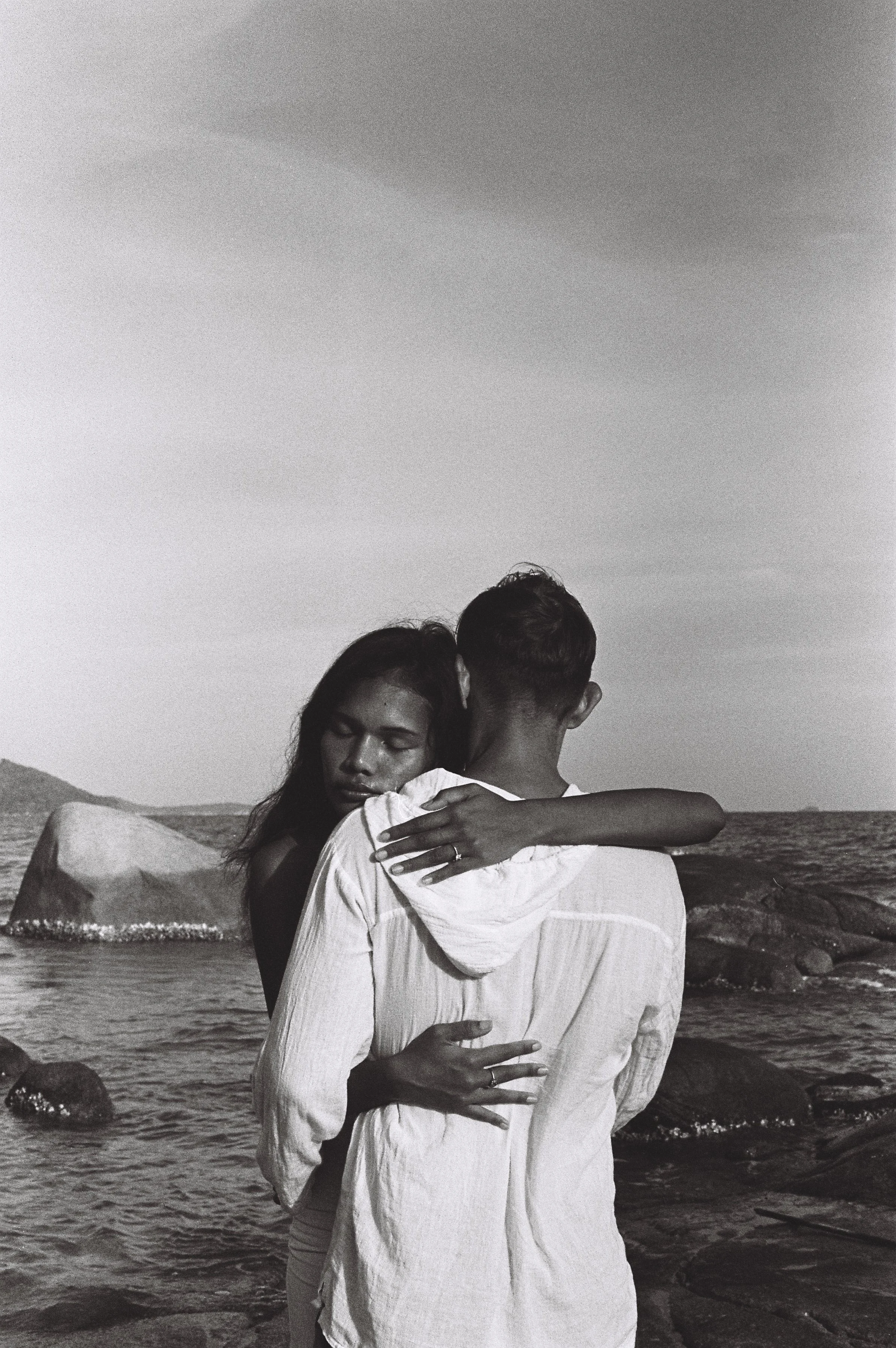 A black and white photo of a couple embracing each other near the shoreline with rocky landscape and ocean in the background.
