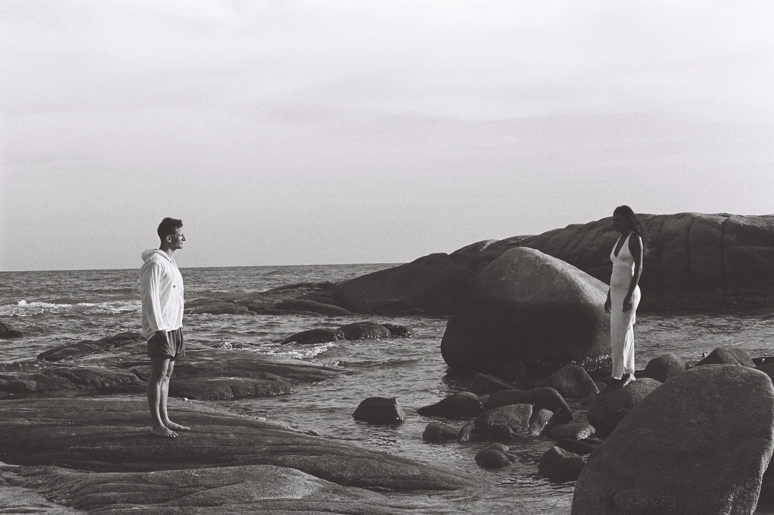 A black-and-white photo of a man and a woman standing on rocks by the ocean. The man is on the left, wearing a hoodie and shorts, looking to the right. The woman is on the right, wearing a dress, looking down, with large rocks behind her. The sky is overcast.
