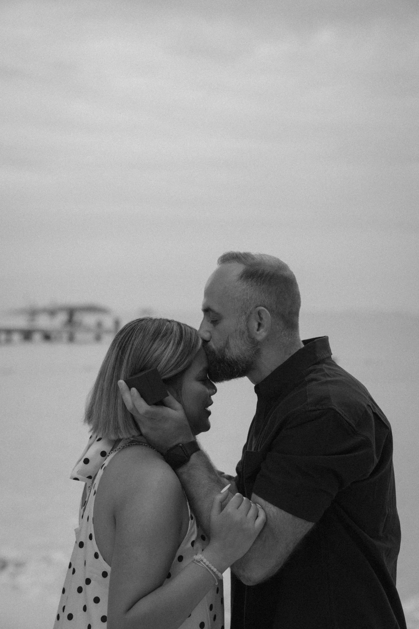 A black and white photo of a couple kissing outdoors, with the woman holding the man's face and the man holding the woman's hand.