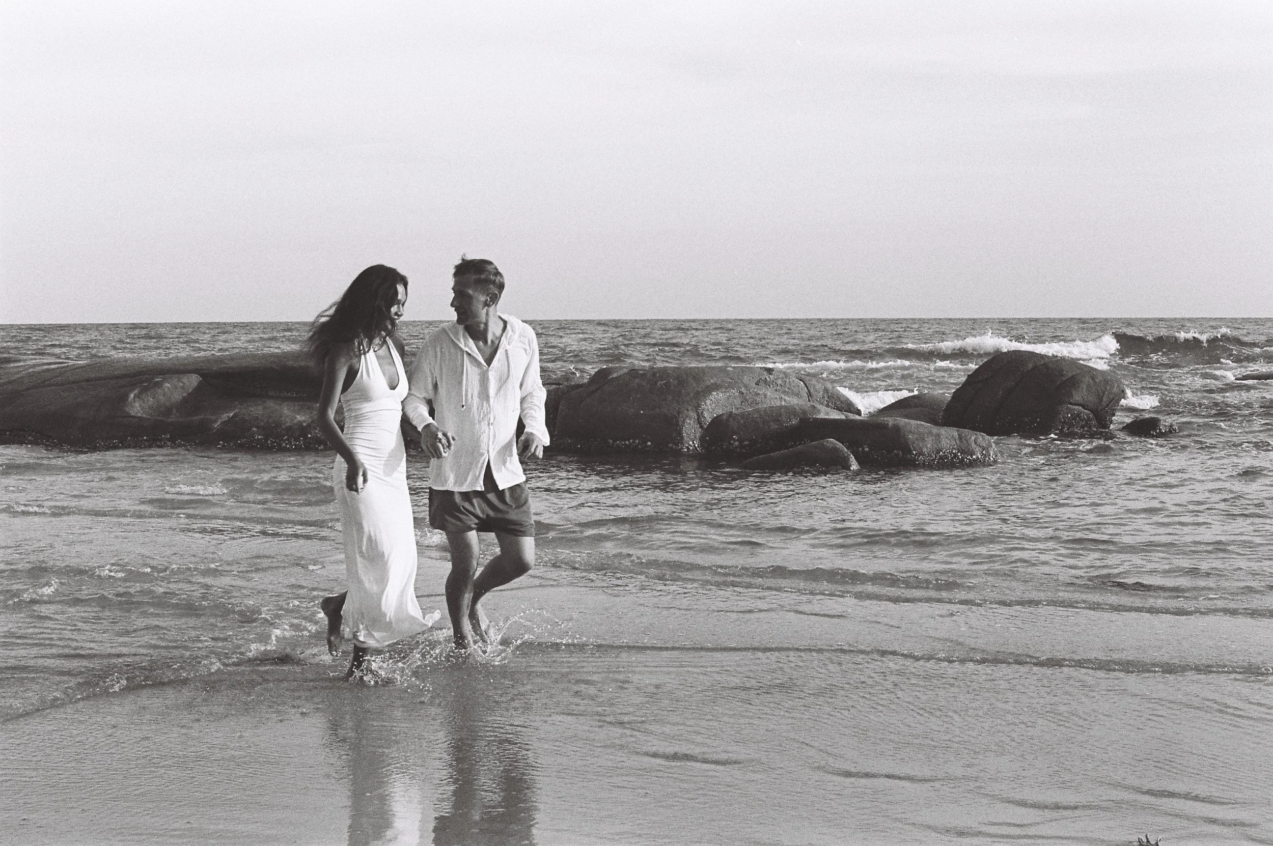 A man and woman holding hands and walking on the beach near rocks and the ocean in black and white.