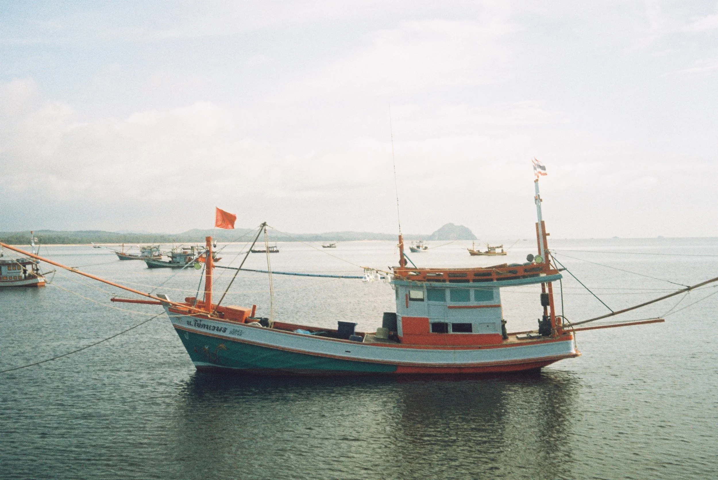 Fishing boats docked on calm water near a harbor during daytime, with cloudy sky and distant land in background.