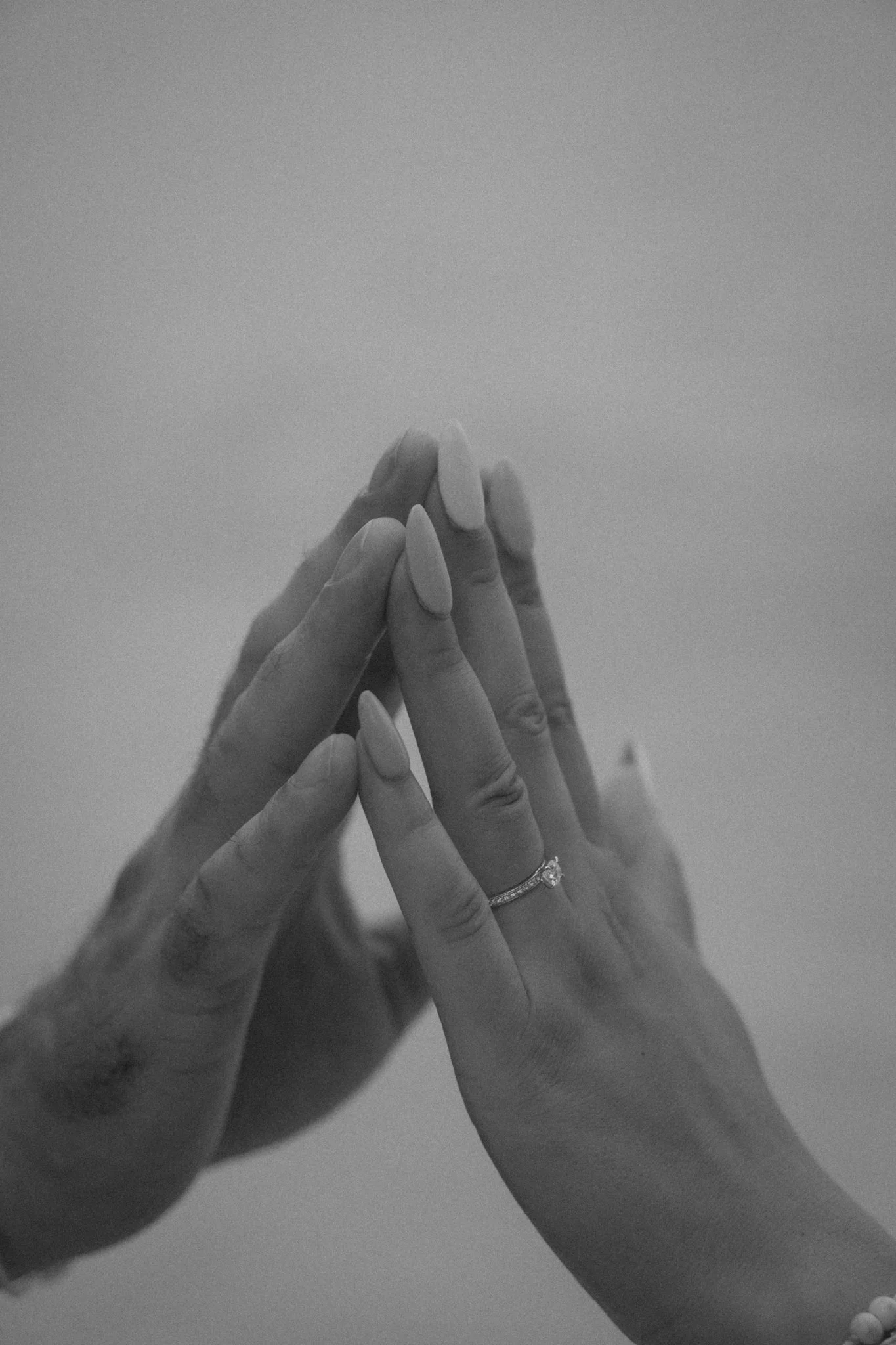 Multiple hands with manicured nails and rings, gently touching each other, against a plain background.