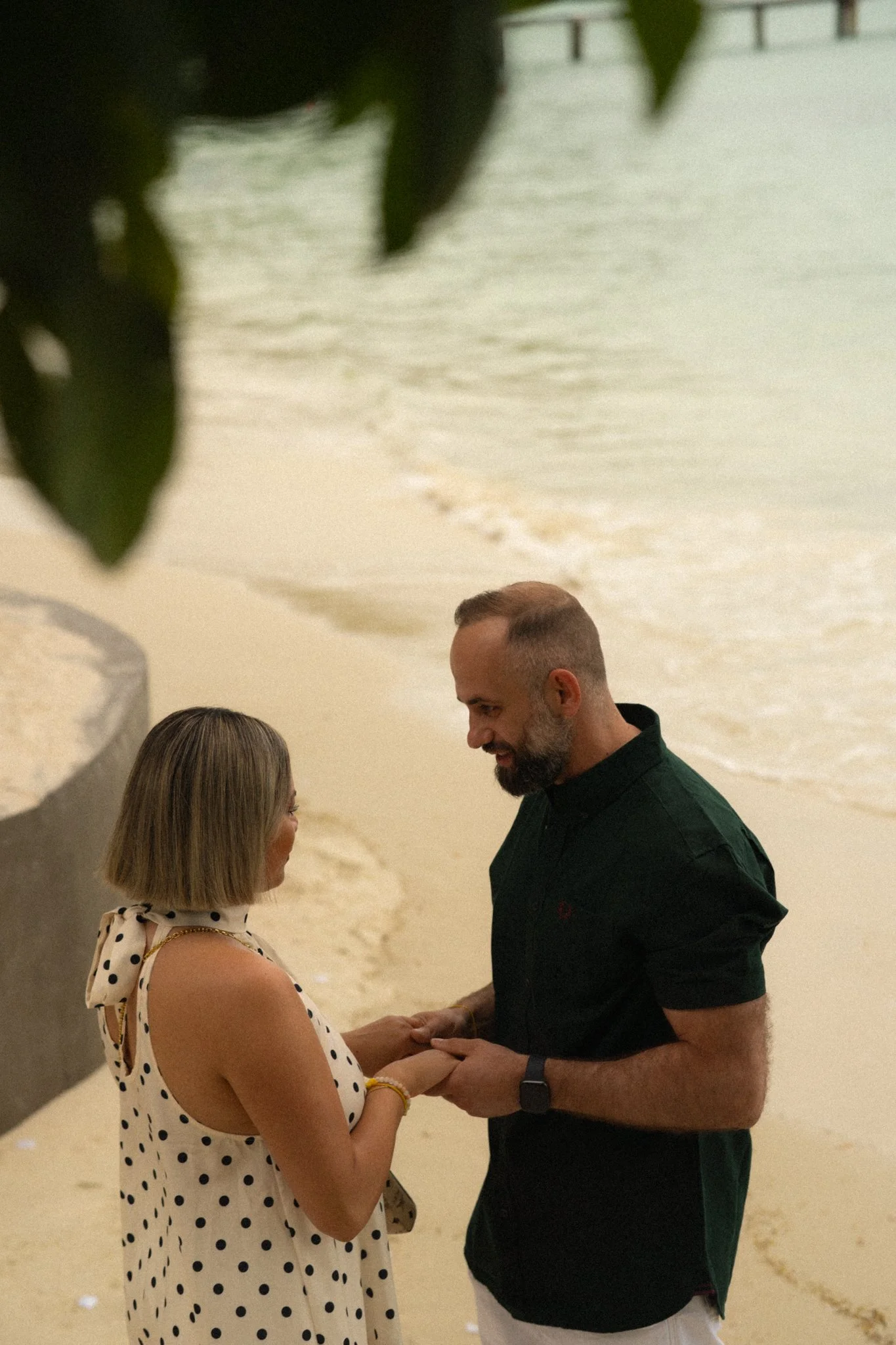 A woman with blonde hair and a polka dot dress and a man with a beard and a black shirt share a moment on a beach during sunset, holding hands and looking into each other's eyes.