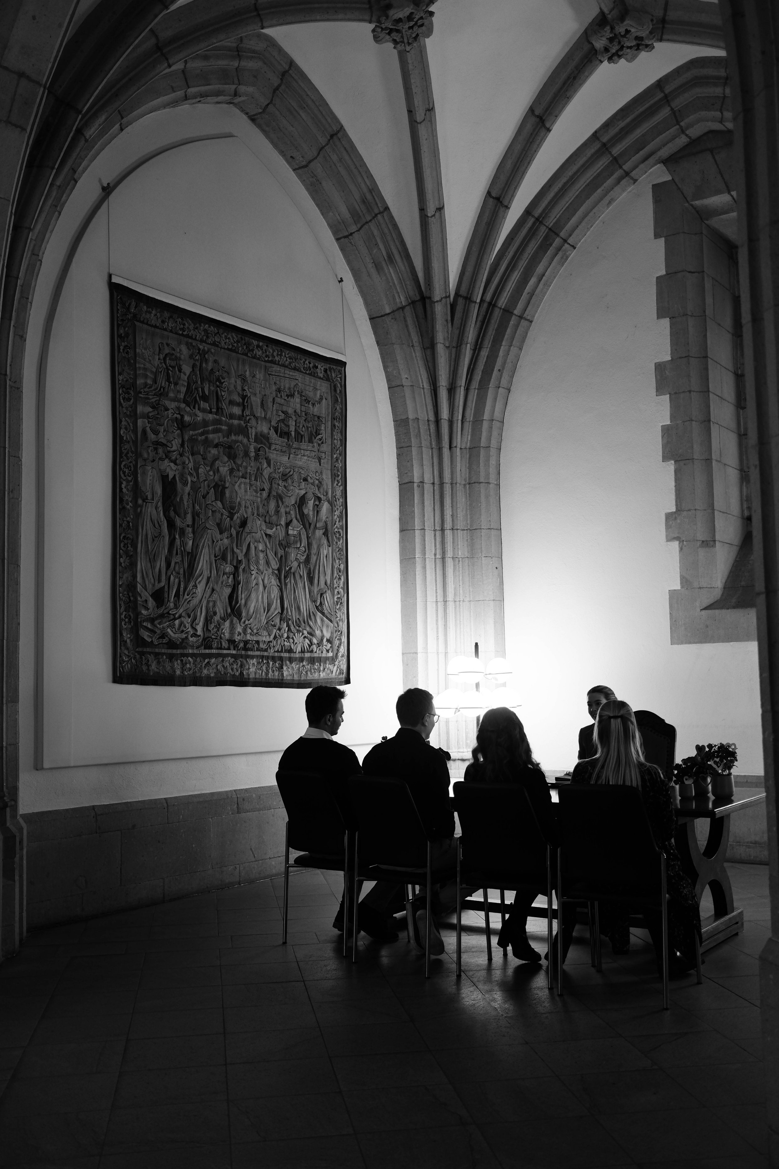 Fünf Personen sitzen an einem Tisch in einem gotischen Raum mit hohen, gewölbten Decken, einer Wand mit einem großen Wandteppich und einer Lampenleuchte zwischen ihnen.