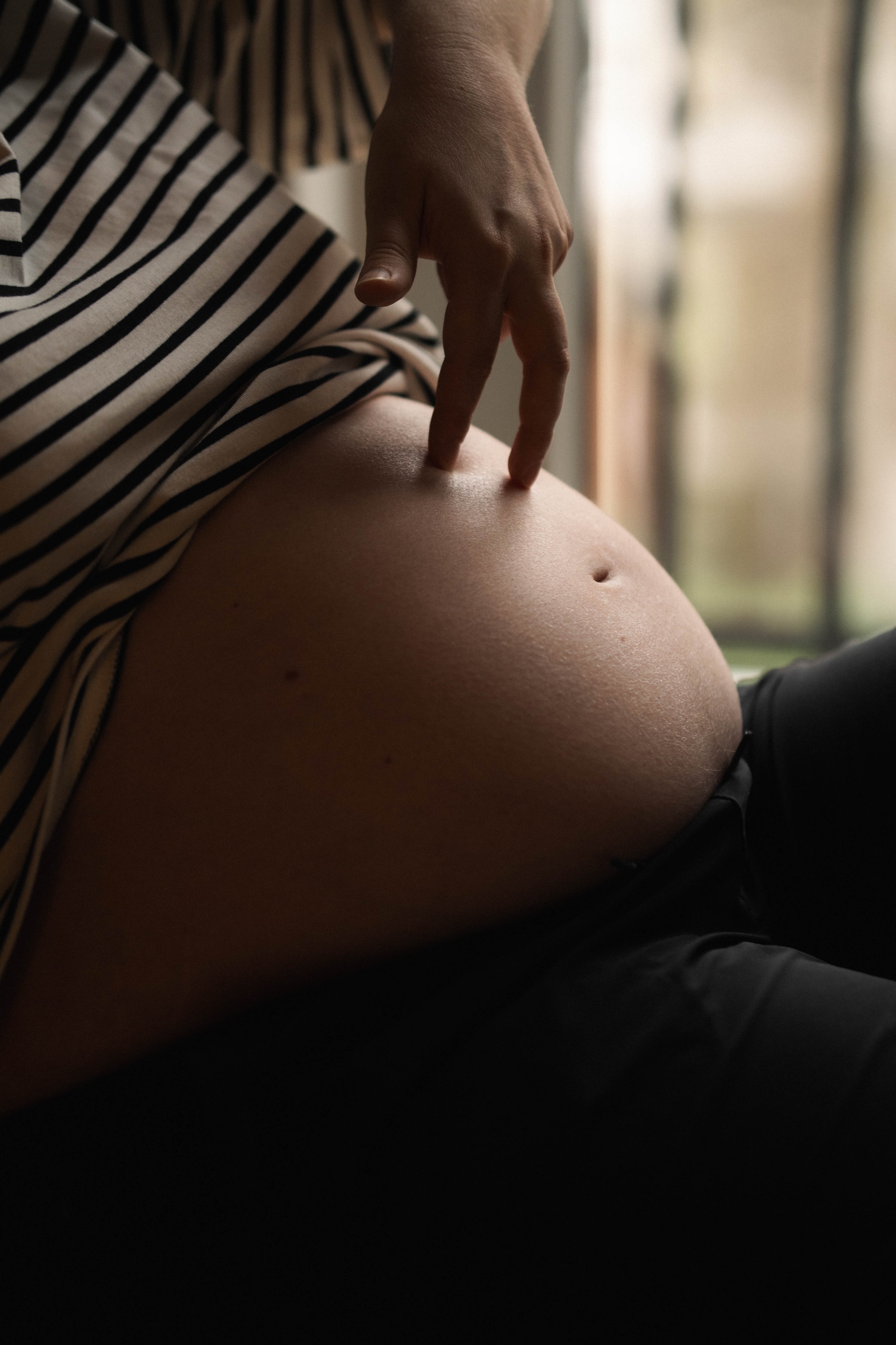 Close-up of a person's pregnant belly with a hand touching it, wearing black pants and a striped shirt, near a window.