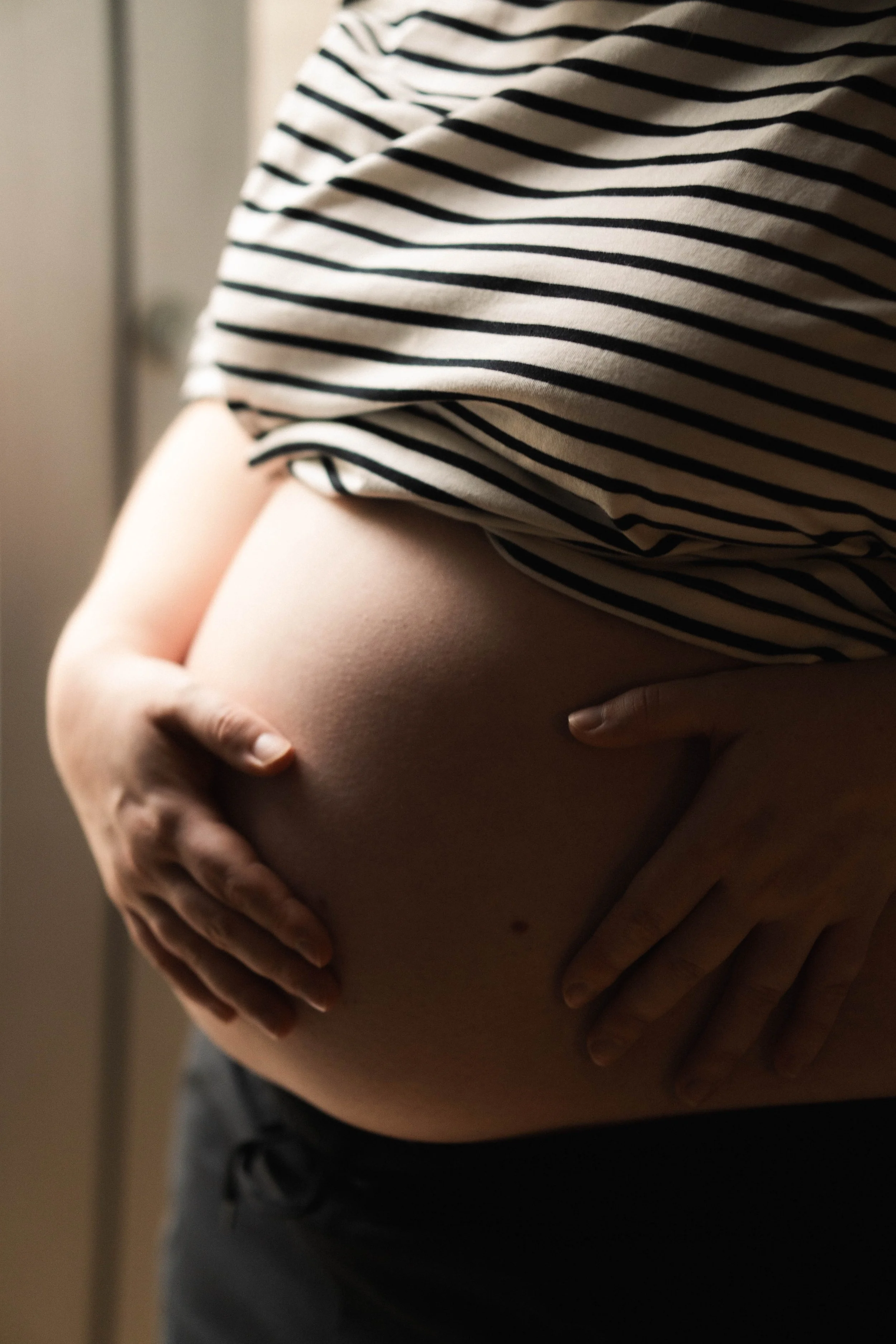 Close-up of a pregnant person cradling their bare belly with both hands, wearing a striped shirt, with the background out of focus.