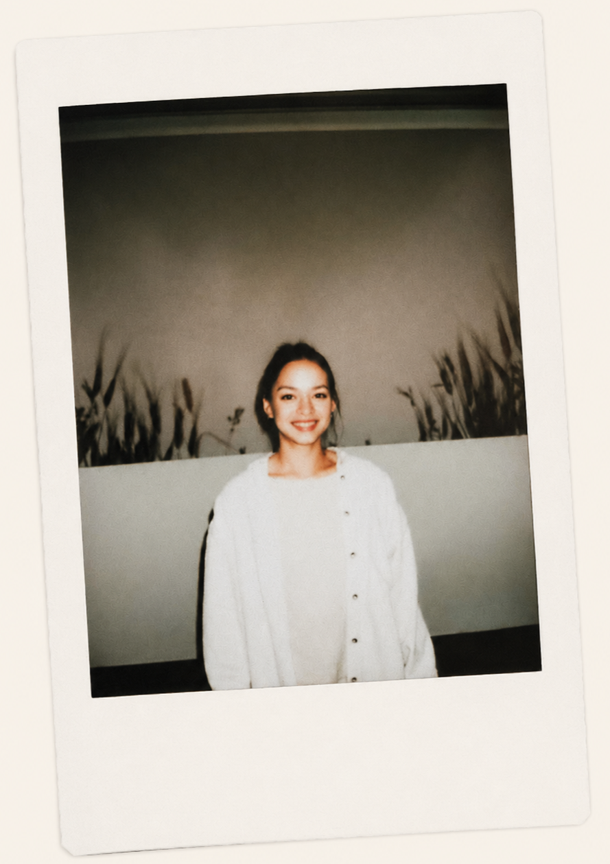 A young woman with dark hair, smiling, wearing a white oversized sweater, standing in front of a plain wall with plant shadows.