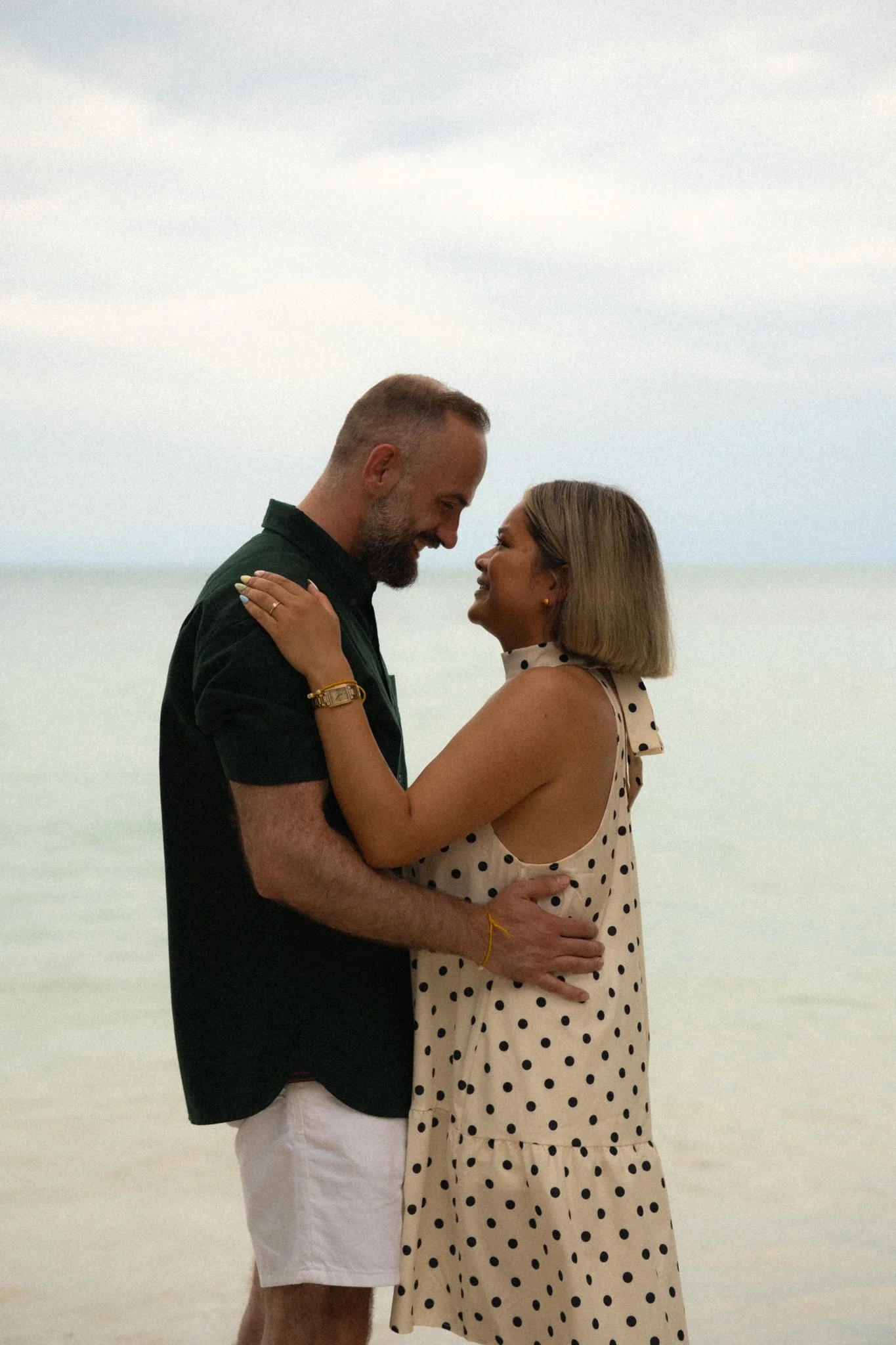 A couple standing close together on a beach, smiling and looking into each other's eyes. The man has a beard and is wearing a black shirt and white shorts. The woman has shoulder-length hair and is wearing a cream-colored dress with black polka dots.