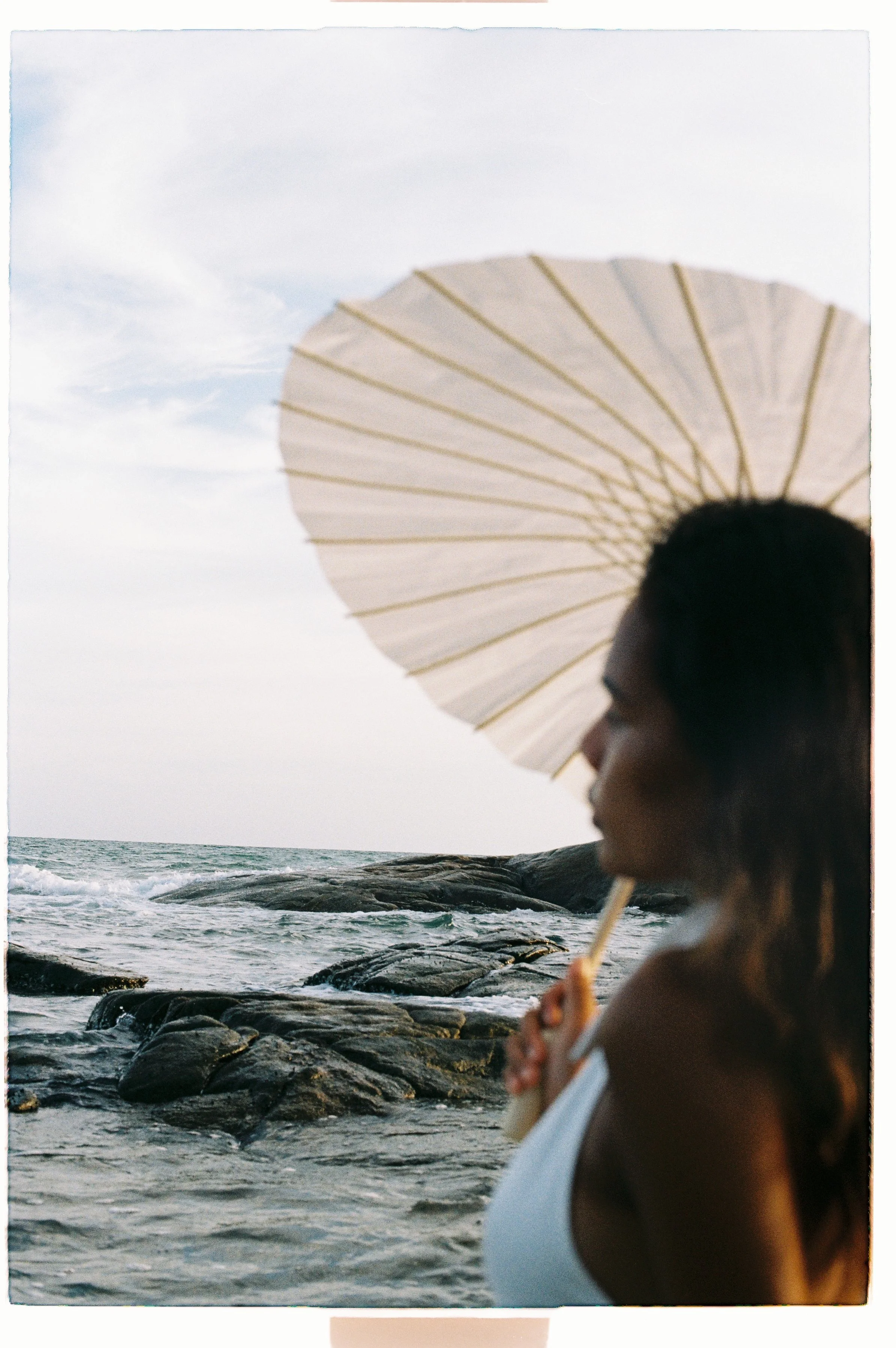A woman with dark hair holding a white parasol at the beach, with waves and rocks in the background, wearing a white sleeveless top.