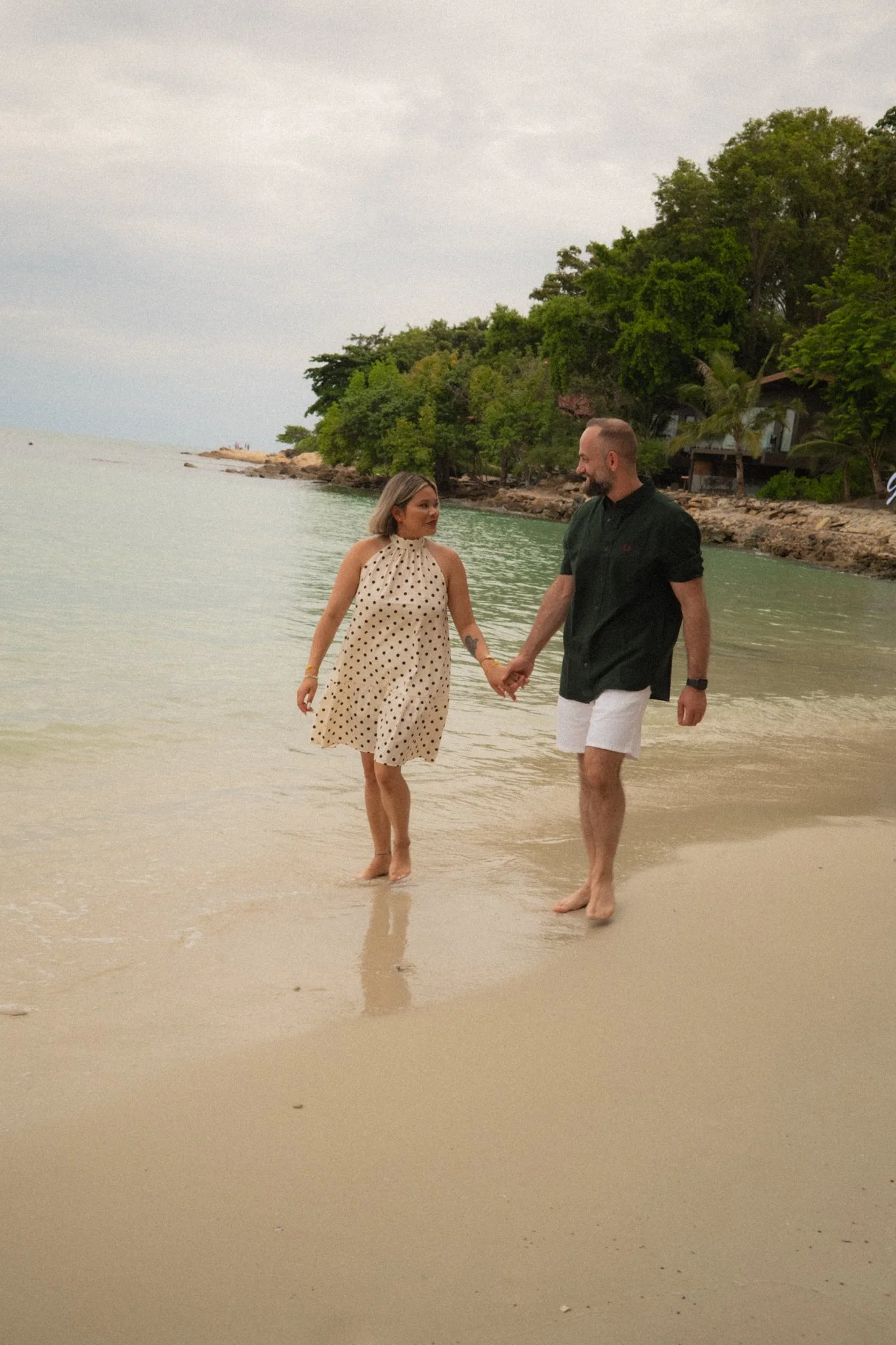 A couple holding hands and walking in shallow ocean water near a beach with greenery and trees in the background.