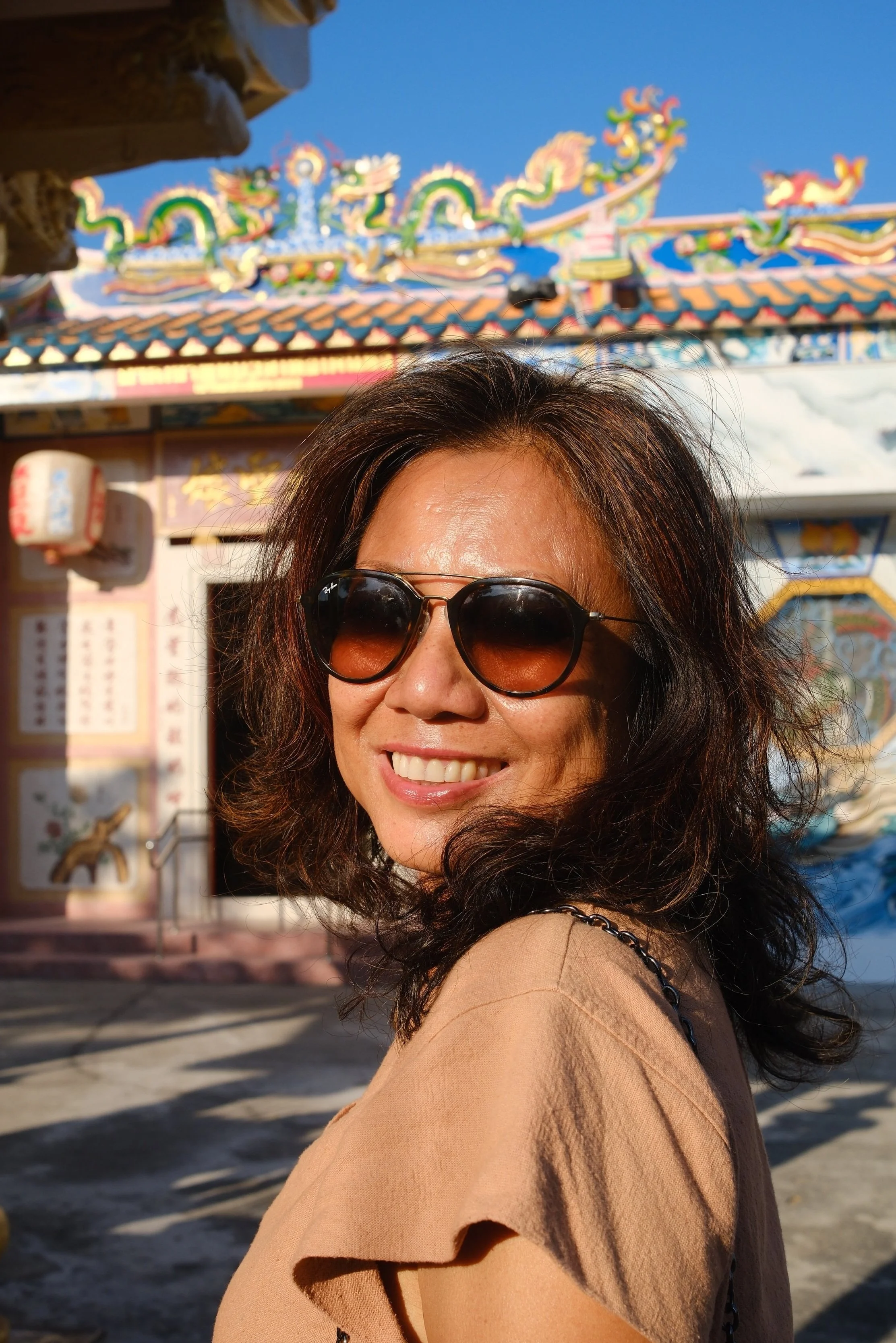 Ein lachende Frau mit schwarzen, lockigen Haaren, Sonnenbrille und beigem Oberteil vor einer bunten, chinesisch inspirierten Tempelanlage bei Sonnenuntergang.