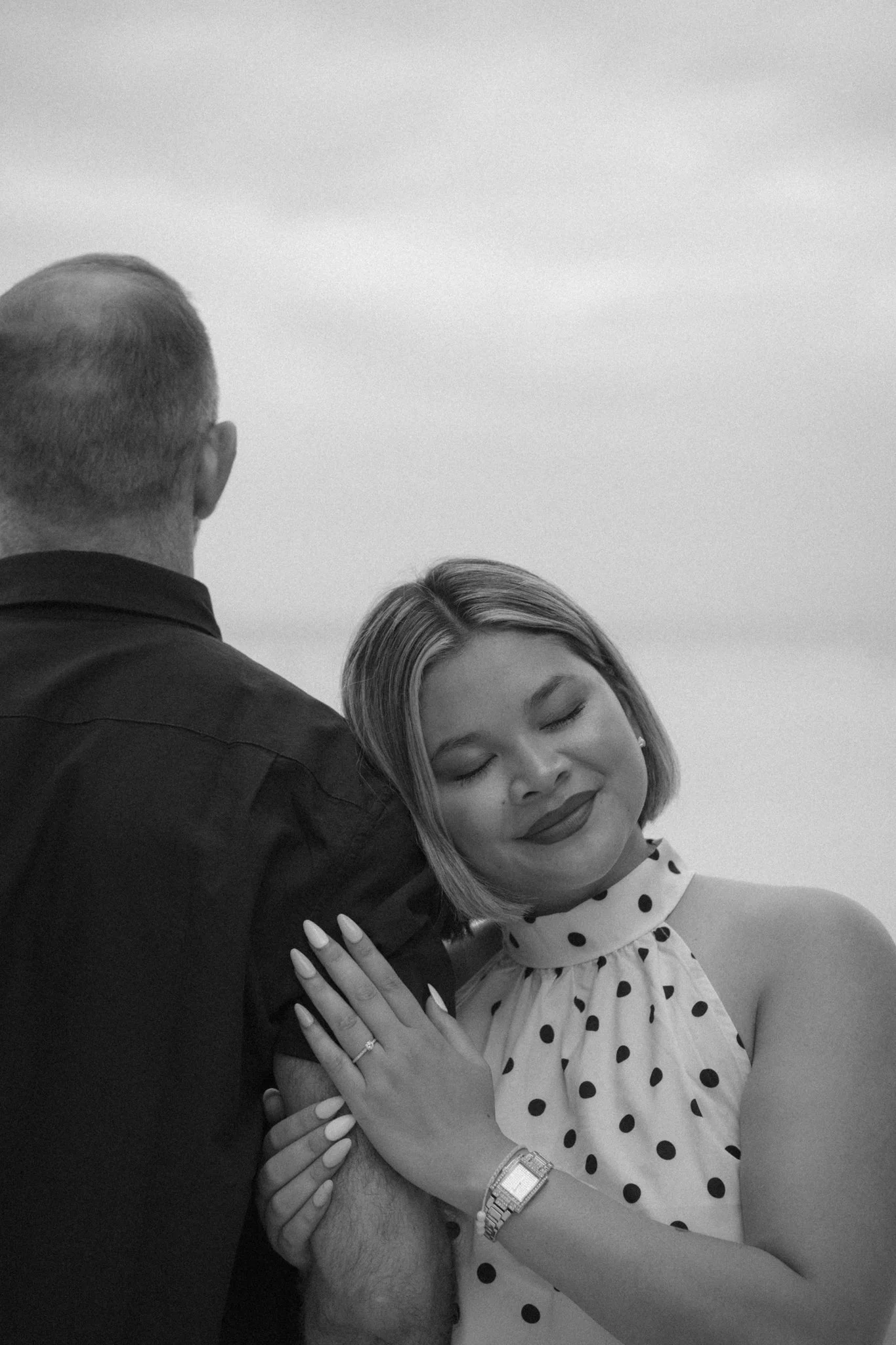 A woman with a bob haircut and a polka dot dress is smiling with her eyes closed, leaning into a man who is facing away from the camera. The man is wearing a dark shirt and a wristwatch. The woman has her hand on the man's chest, showing an engagemen