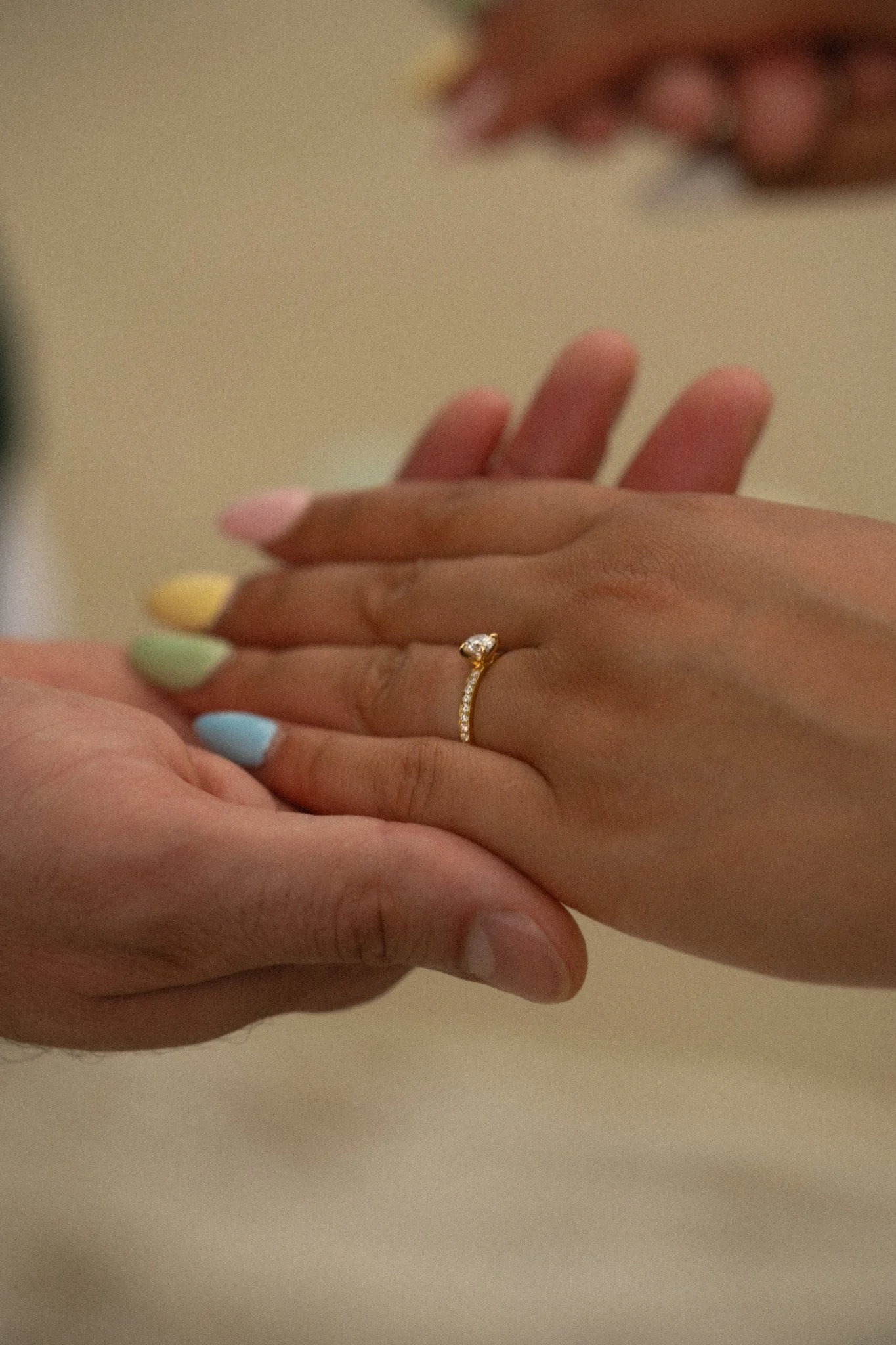 Close-up of two hands, one with an engagement ring with a diamond, gently touching each other against a beige background.