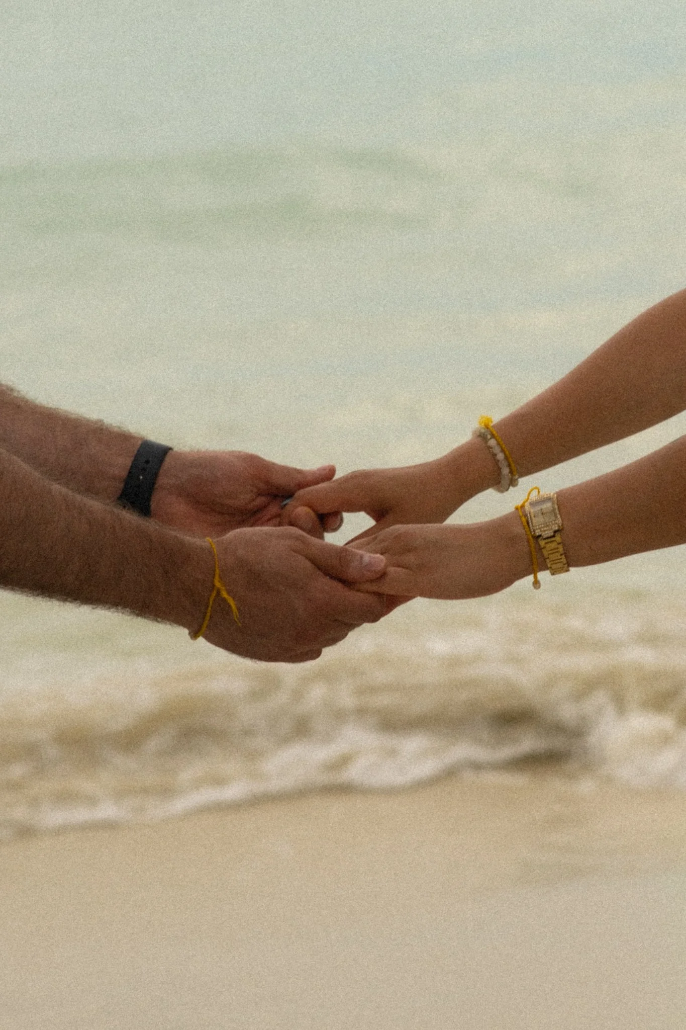 Two people holding hands on the beach, wearing bracelets and watches, with the ocean in the background.