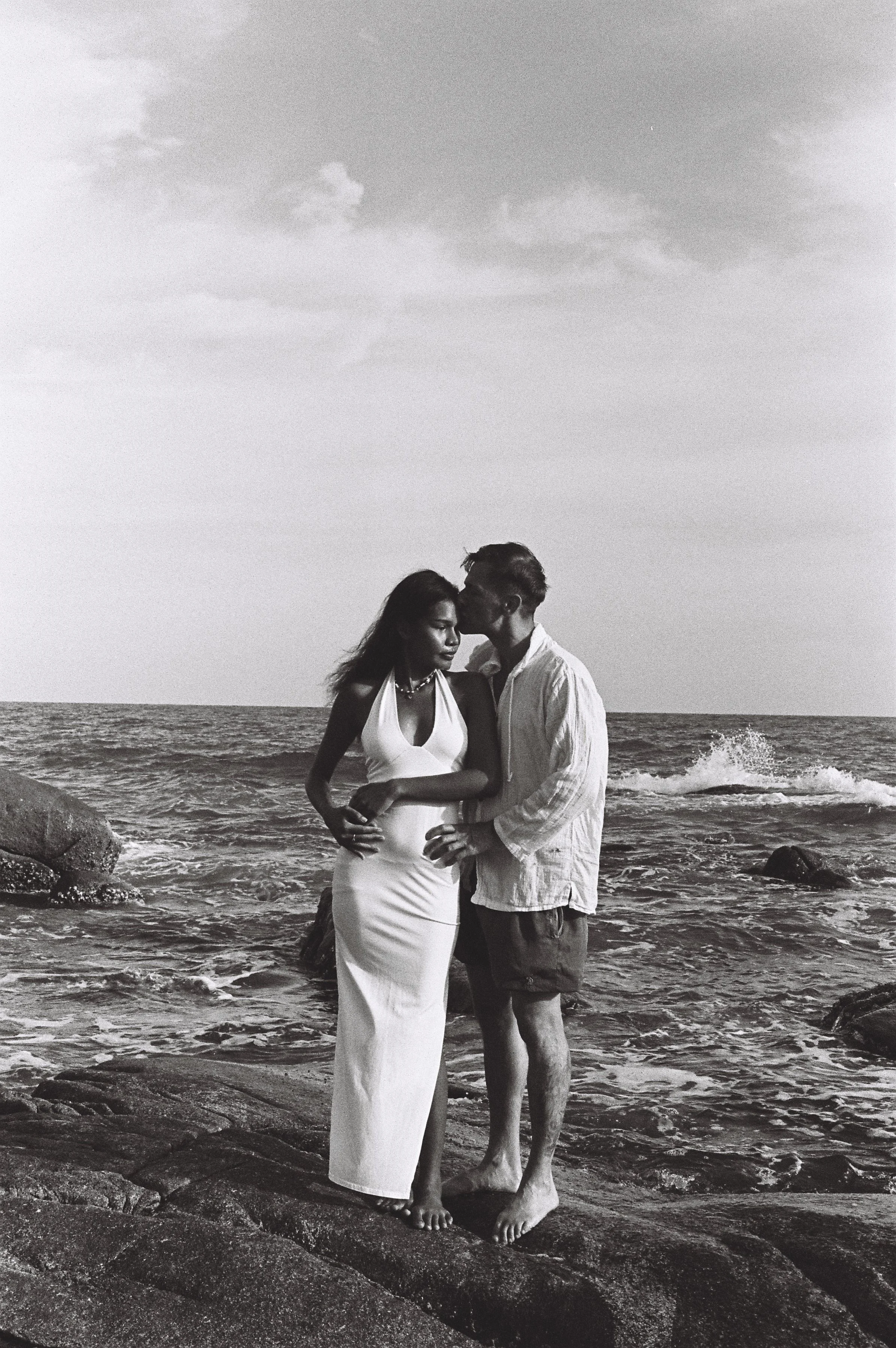 A couple stands on rocks by the ocean, embracing each other, with waves in the background and a cloudy sky overhead.