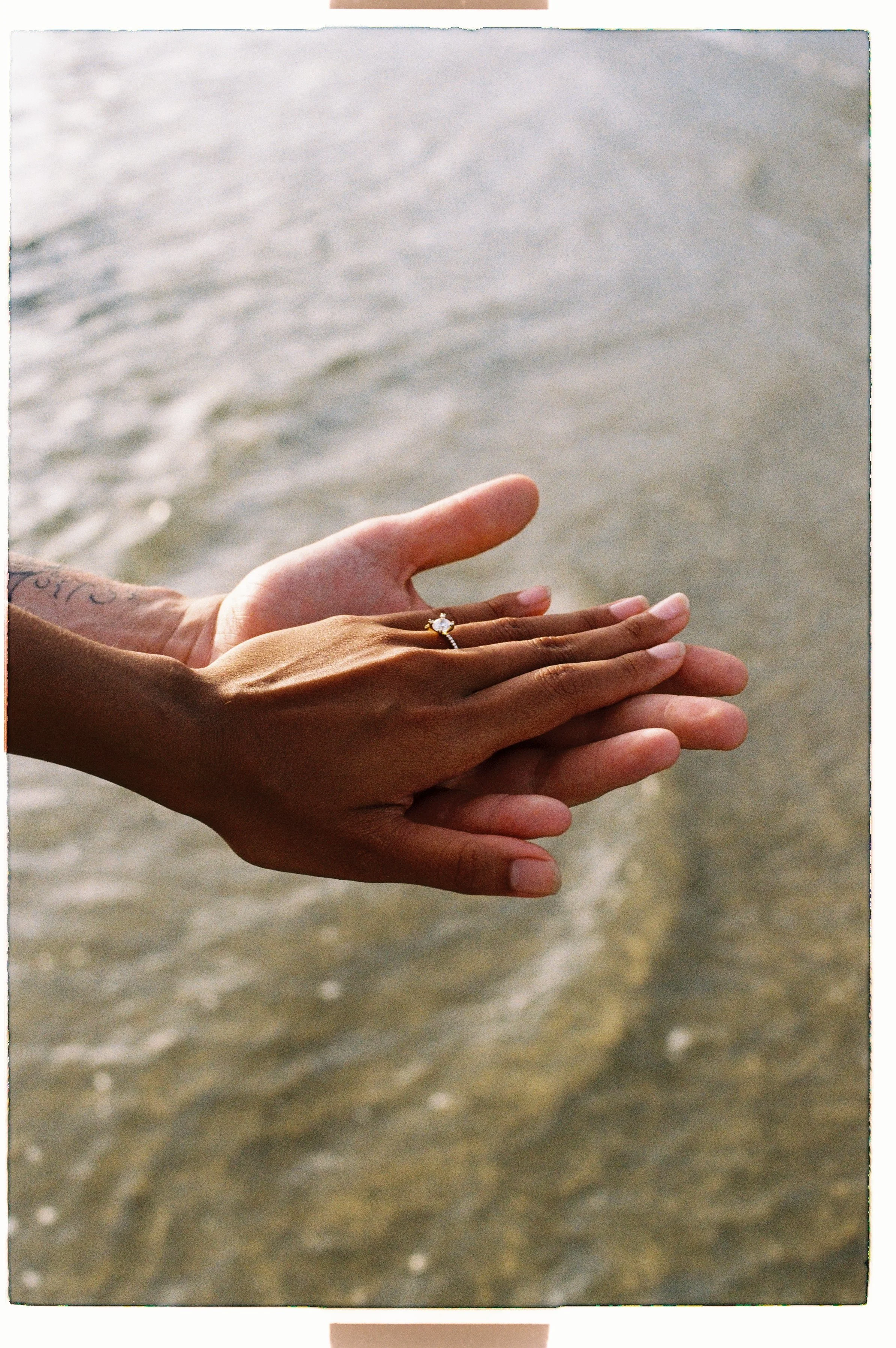 Close-up of two hands with different skin tones touching, with water in the background, and one hand is wearing an engagement ring with a prominent diamond.