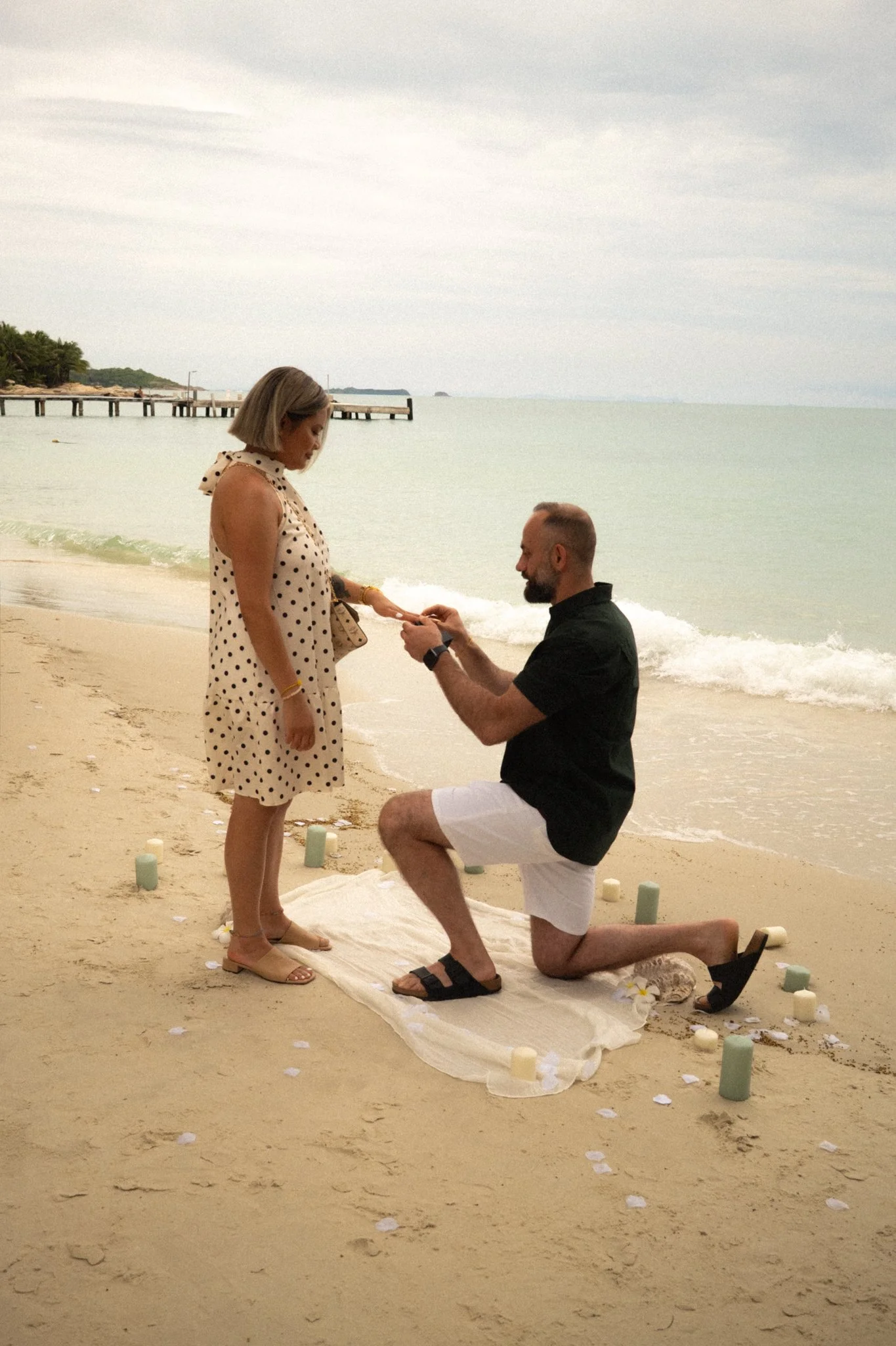 A couple is on a beach during a proposal. The man is kneeling on one knee, holding a ring, and proposing to the woman, who is looking down at him. The scene is decorated with candles and flower petals, with the ocean and pier in the background.