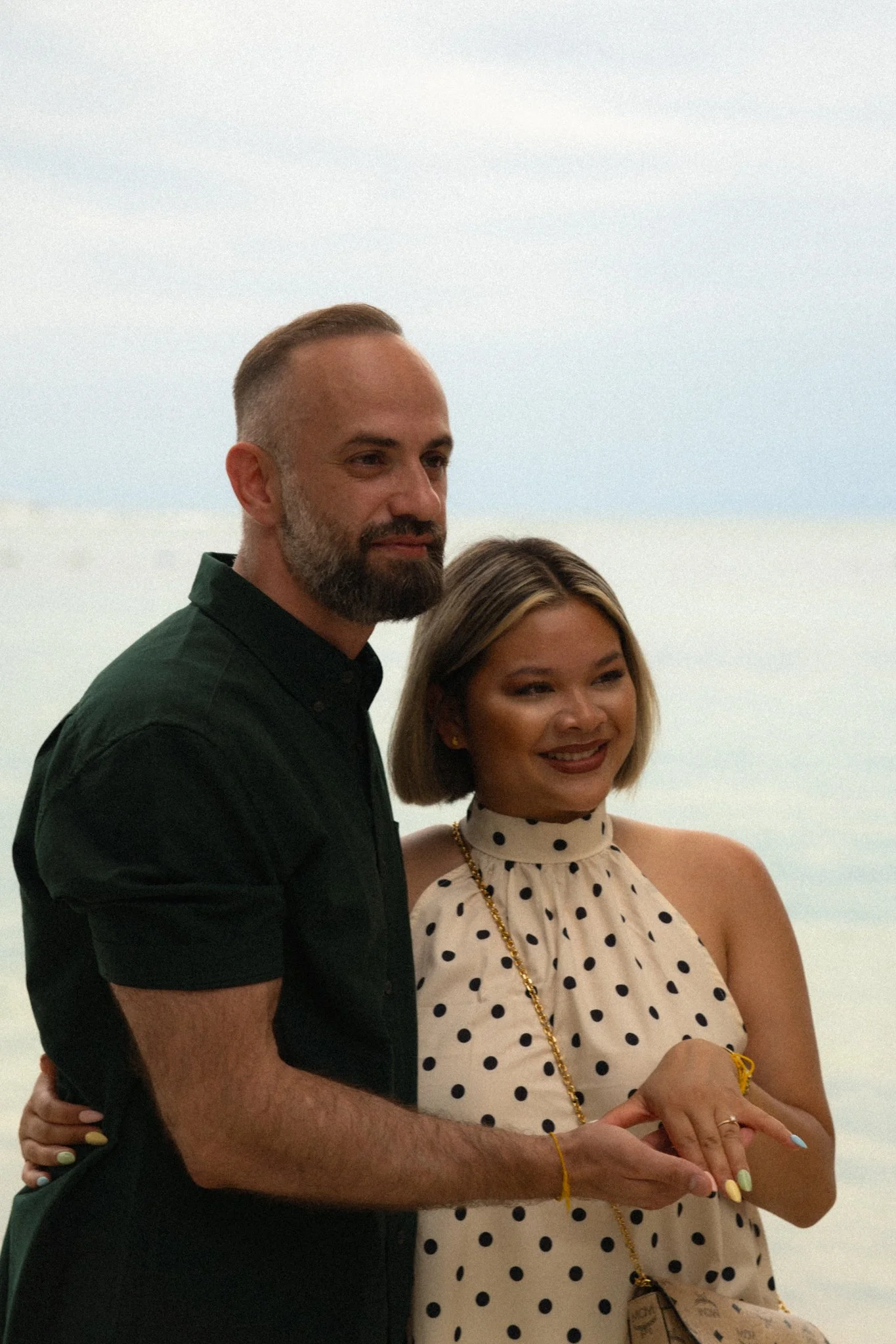 A man with a beard in a black shirt and a woman in a polka dot dress are posing closely together outdoors against a cloudy sky, with the woman's arm around the man's waist.