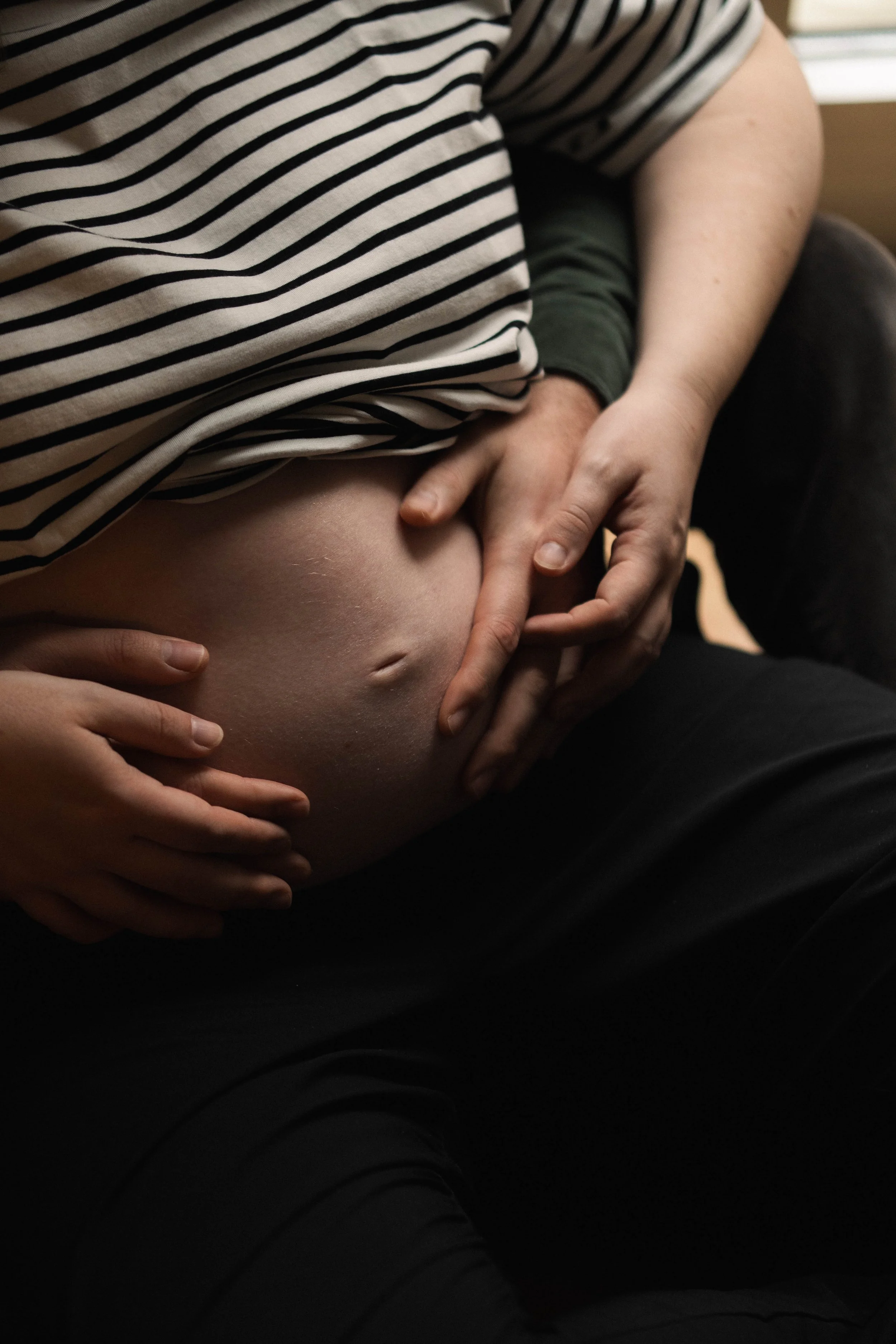 A close-up of a pregnant person's belly with hands gently cradling it, wearing a striped shirt and black pants.