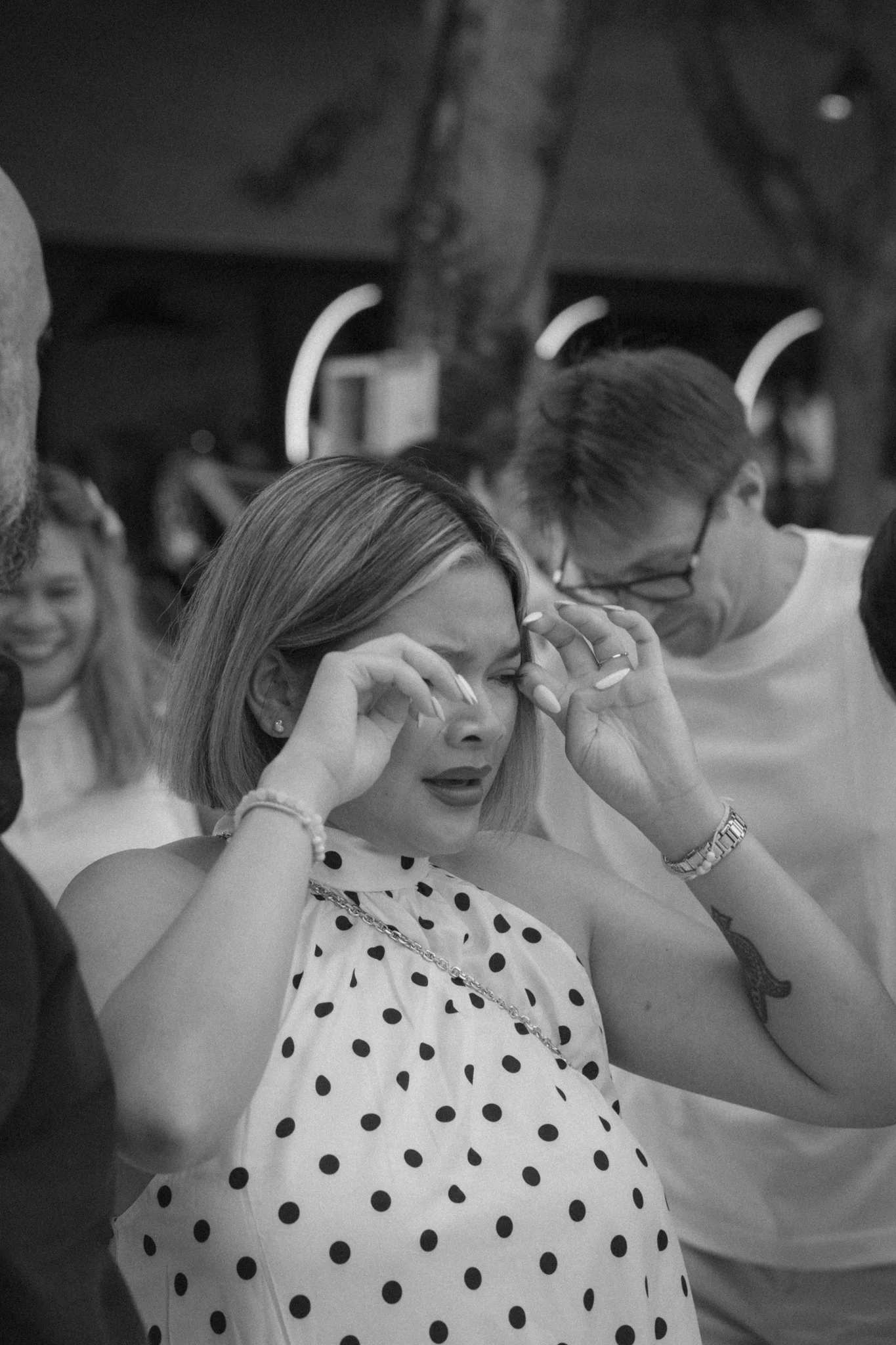 A woman with shoulder-length hair wearing a polka dot dress adjusts her glasses, surrounded by people in a social setting.