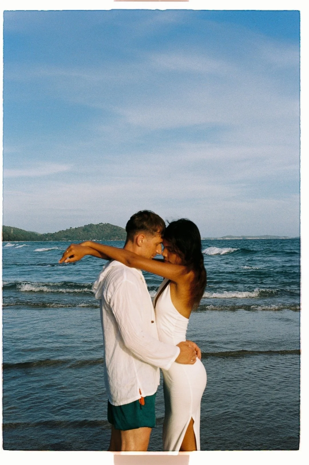 A couple embracing on the beach, standing close with foreheads touching, in front of the ocean with small waves and distant hills under a blue sky.