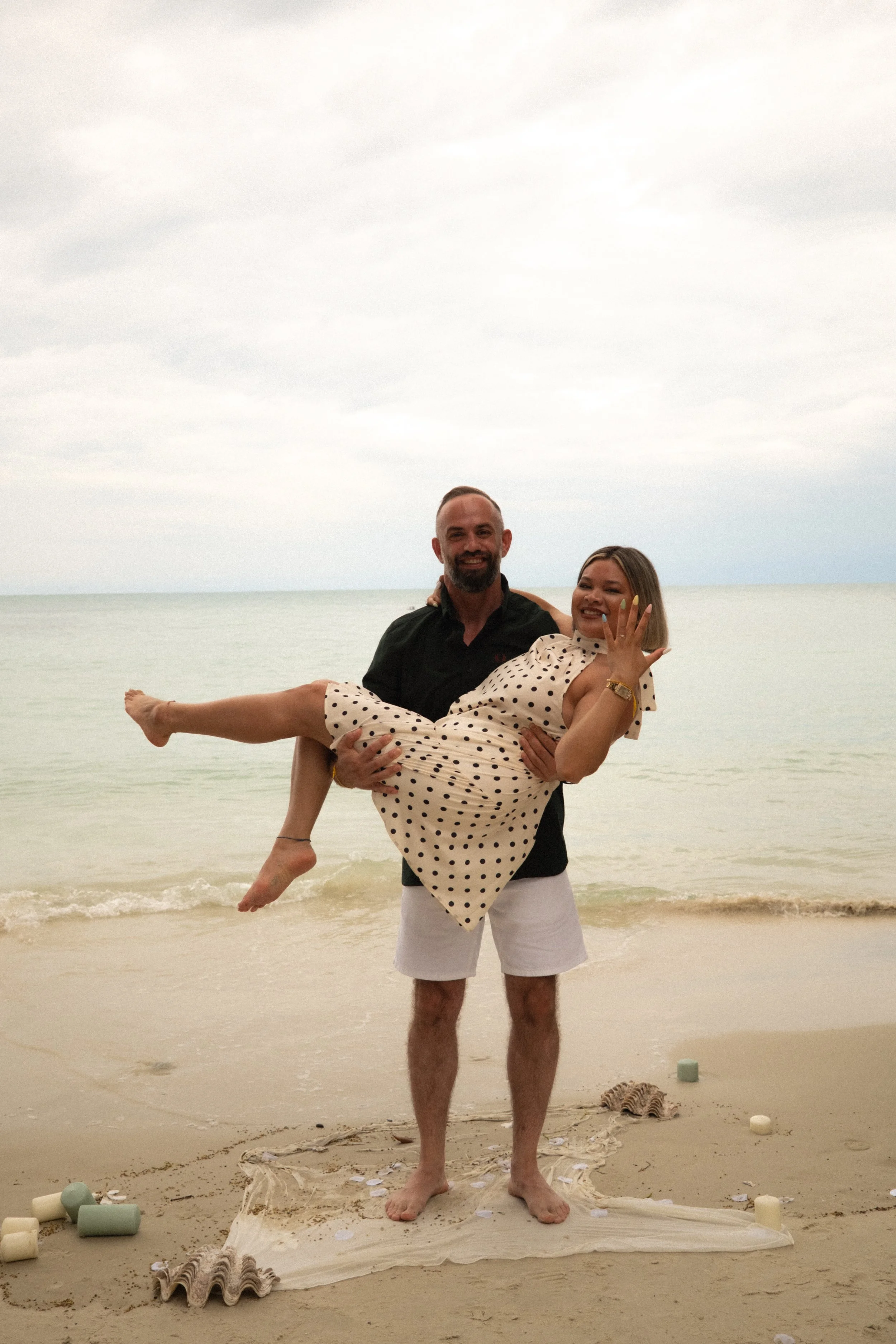 A man in white shorts holds a woman in a polka dot dress in his arms on a beach, with the ocean in the background and a cloudy sky overhead.