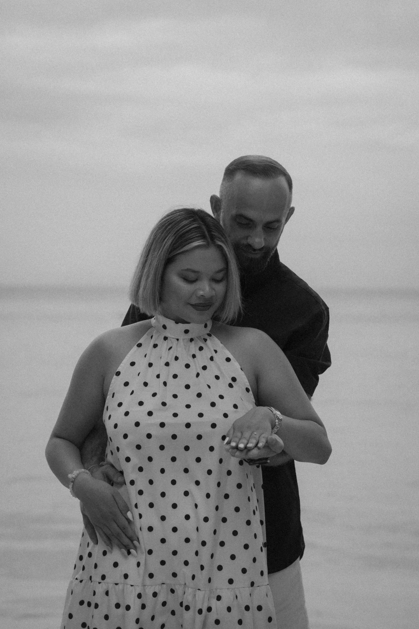 A black and white photo of a woman and a man embracing on a beach. The woman is wearing a polka dot dress and the man is wearing a dark shirt. They are looking down at the woman's arm, which the man is holding.