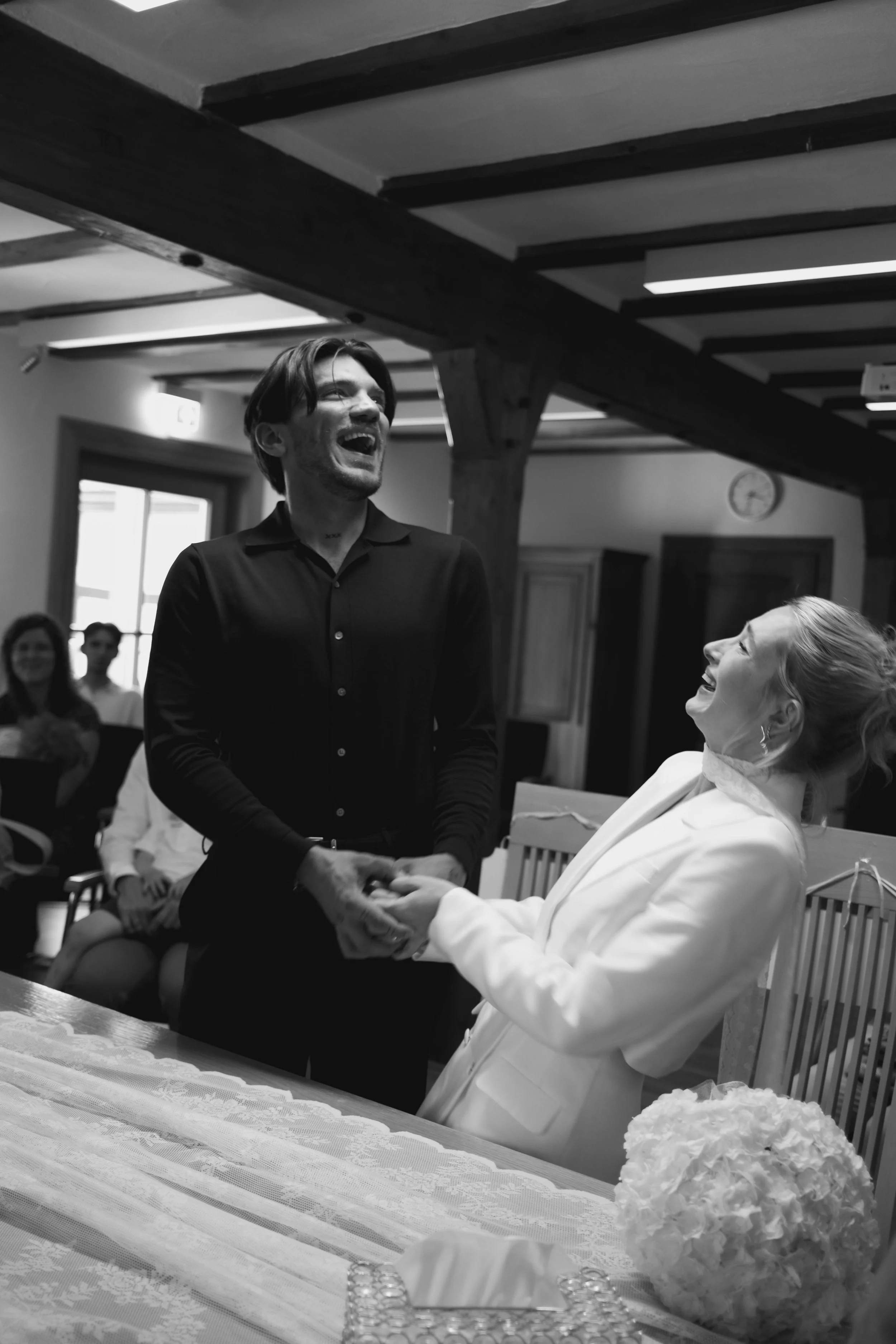 A man in a black shirt and a woman in a white blazer are standing and holding hands, smiling, in a room with a group of people sitting in the background. The room has wooden beams on the ceiling and a window, and there is a floral centerpiece on the table in front of them.