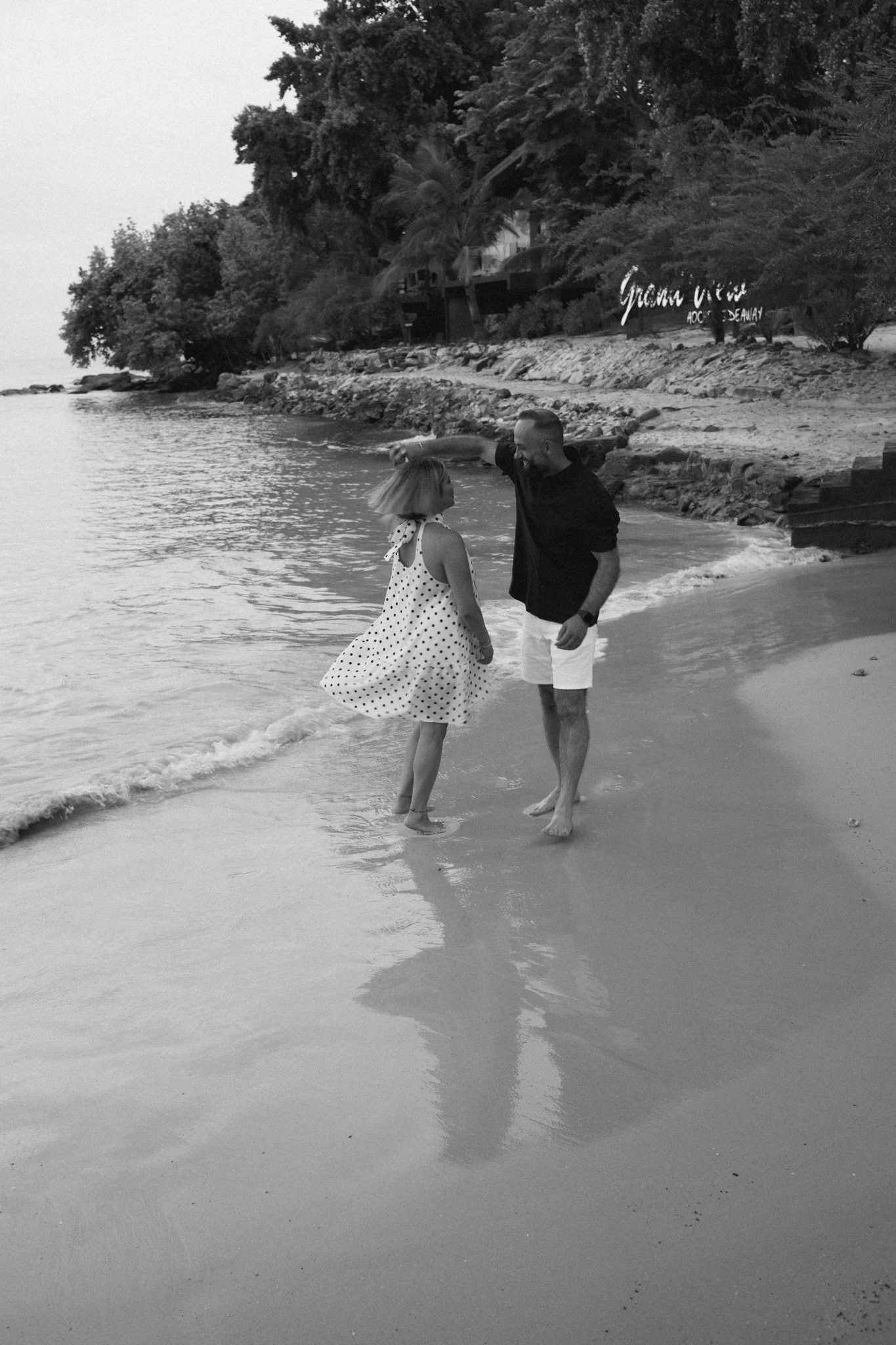 A man and woman dancing on the beach near the water, with trees and a sign in the background. It is in black and white.