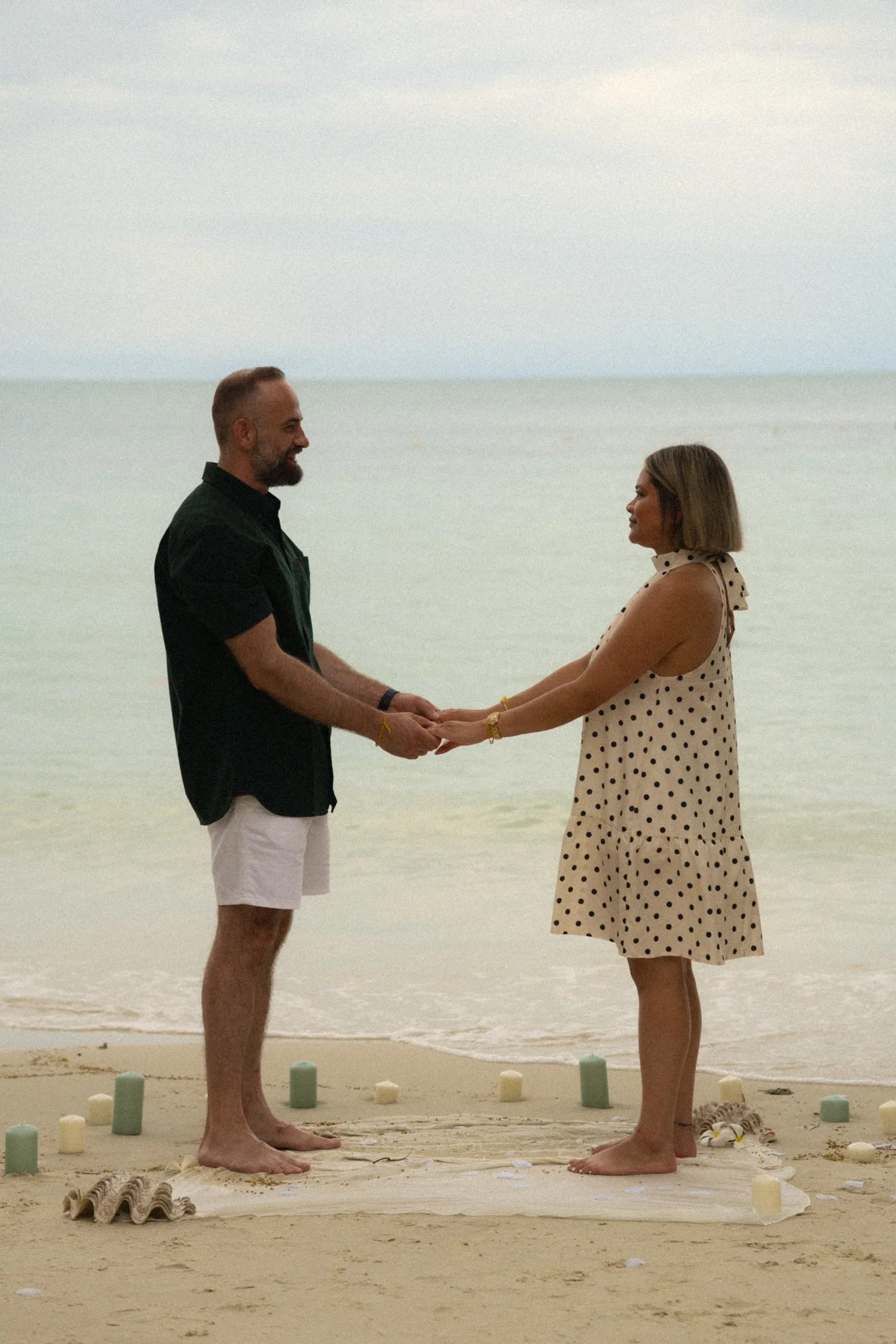 A couple holding hands on a beach, facing each other and smiling, with the ocean in the background. The man has a beard and is wearing a black shirt and white shorts. The woman has shoulder-length hair and is wearing a polka dot dress. There are smal