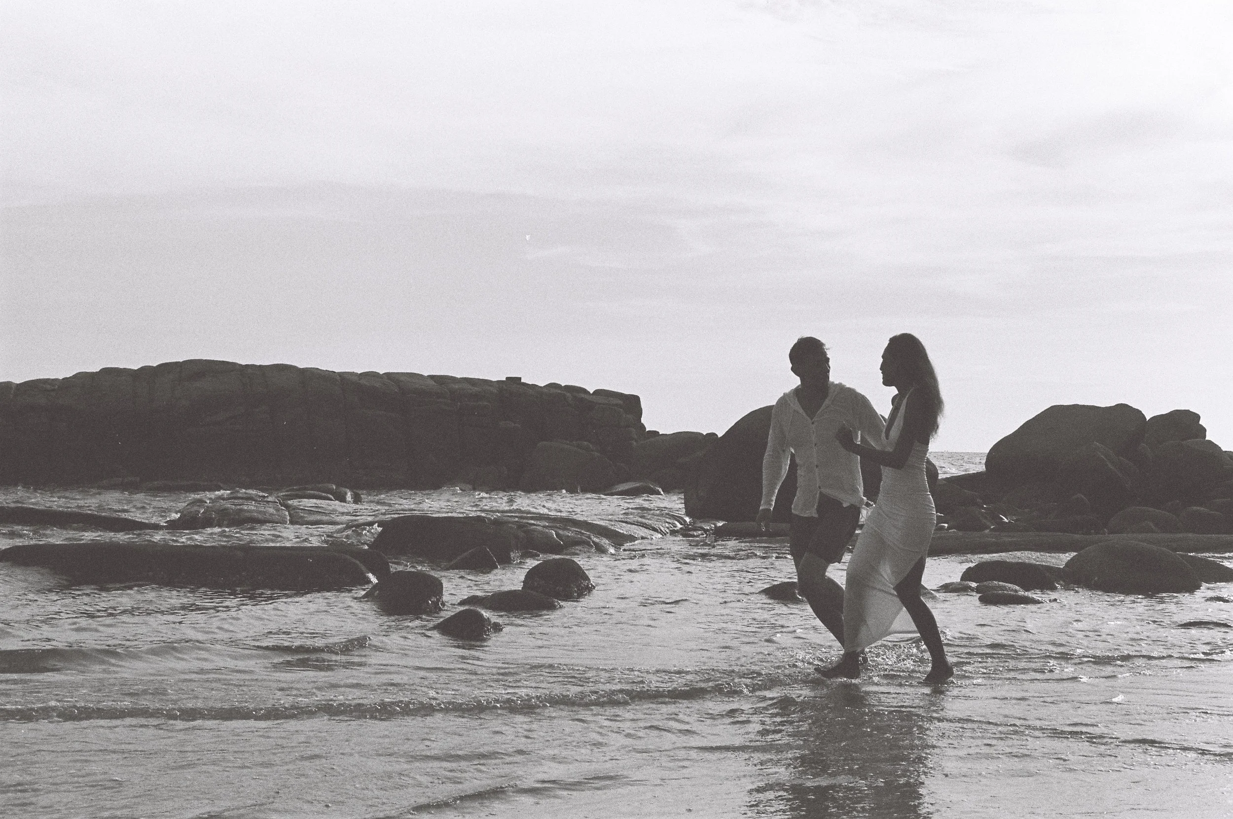 Two people walk along a rocky beach during sunset or dusk, silhouetted against the sky, with large rocks and the ocean in the background.