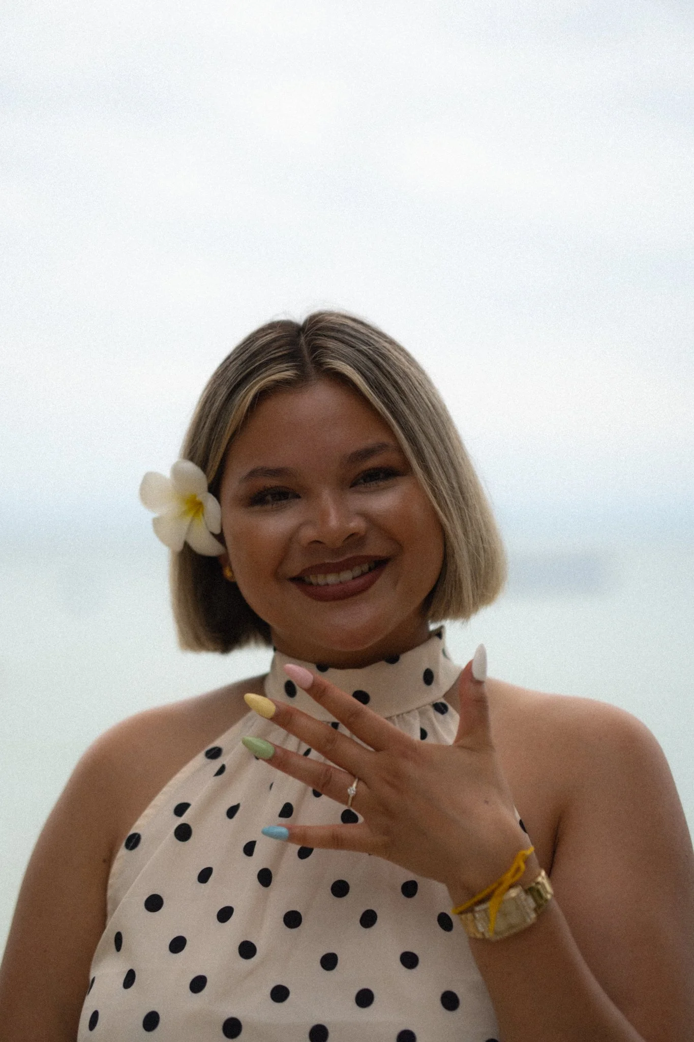 Close-up of a woman smiling, with a white flower in her hair, wearing a polka dot dress, showing her hand with pastel-colored nails, a ring, and a gold watch, against a cloudy sky background.