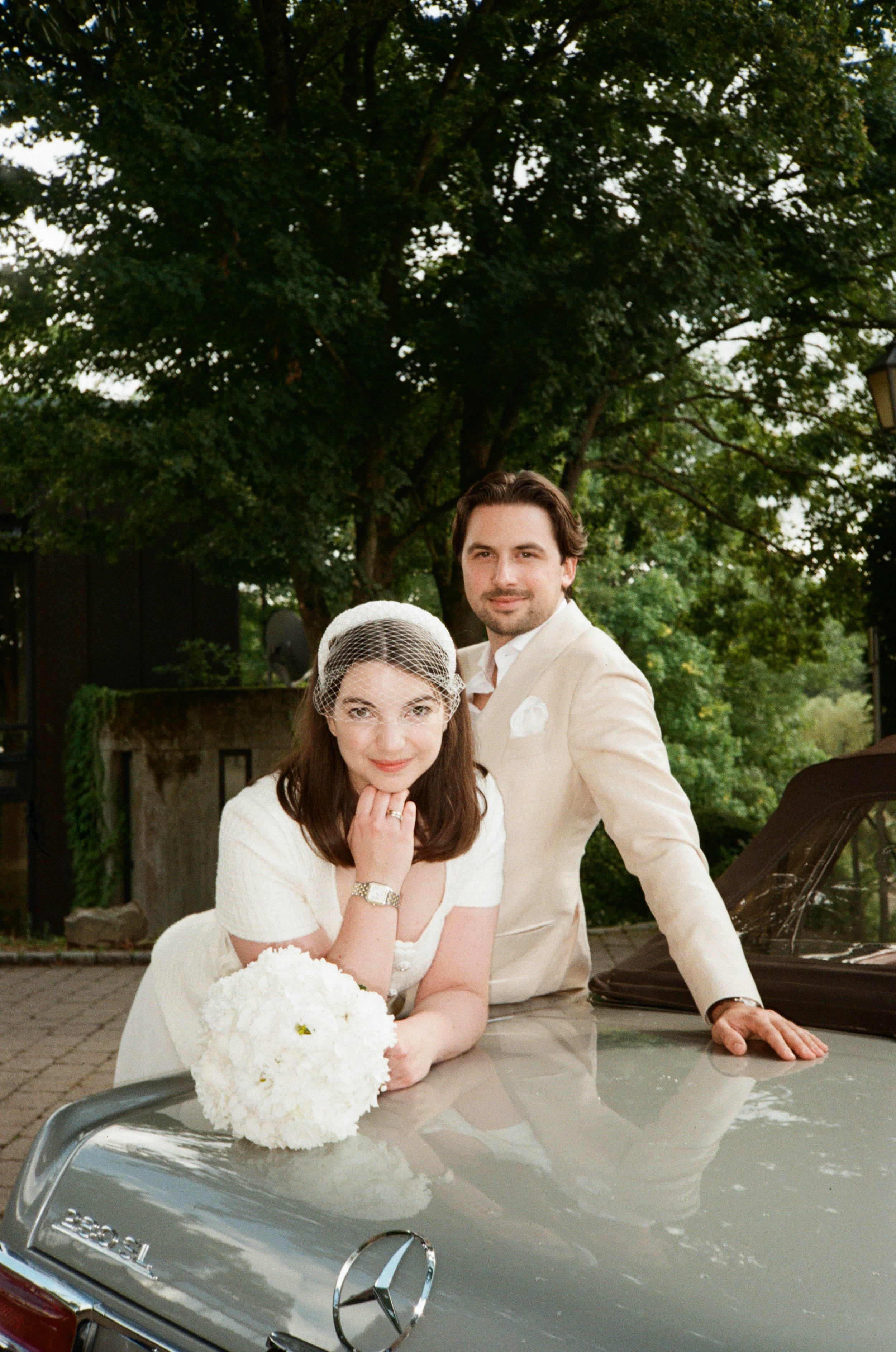 A bride in a white dress with a bouquet of white flowers and a veil, and a groom in a beige suit, are posing outdoors near a silver Mercedes-Benz car, with a large green leafy tree in the background. Wedding Shooting on film.