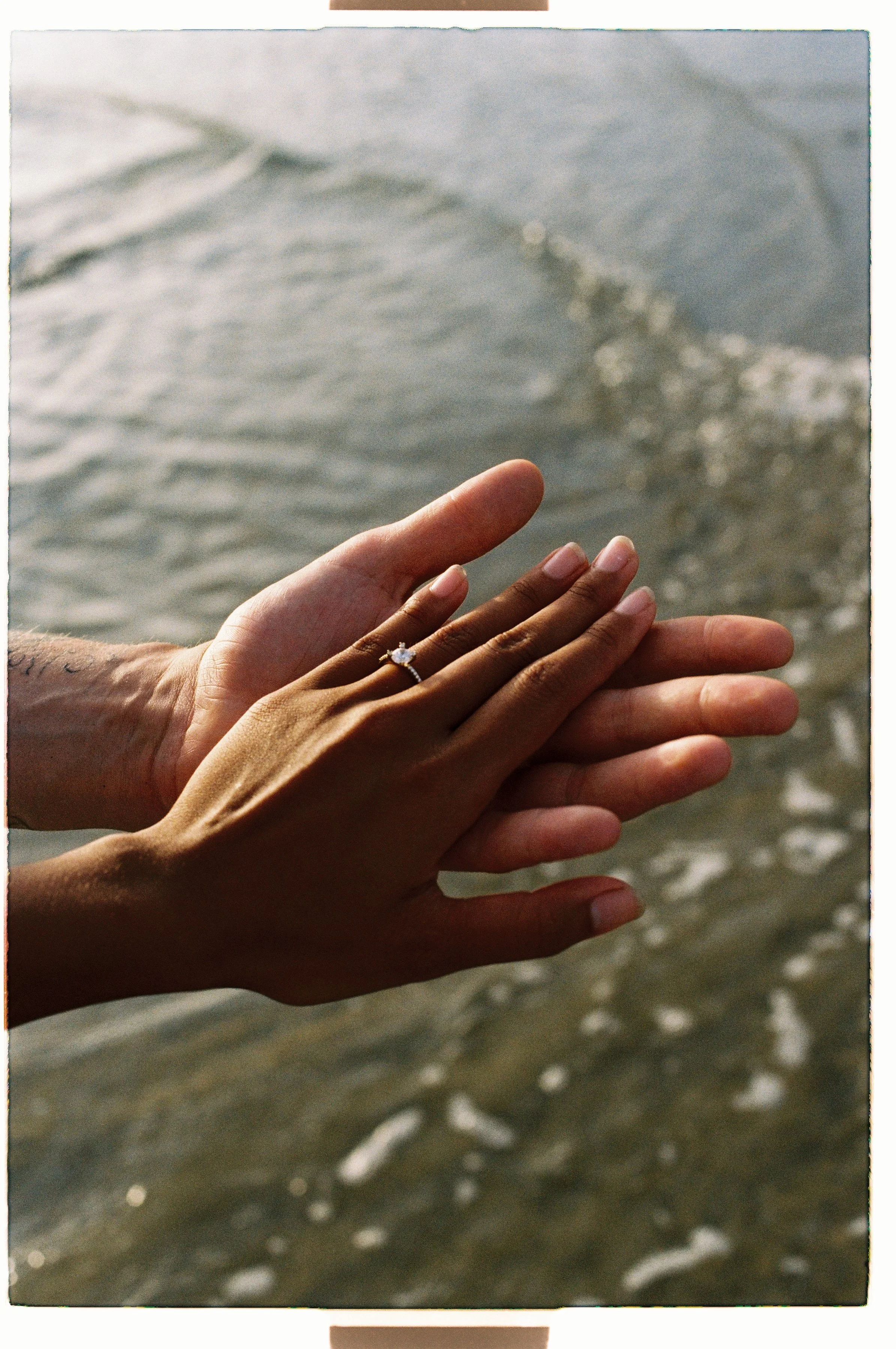 Close-up of two hands, one with a diamond ring on the ring finger, reaching towards the ocean with gentle waves in the background.